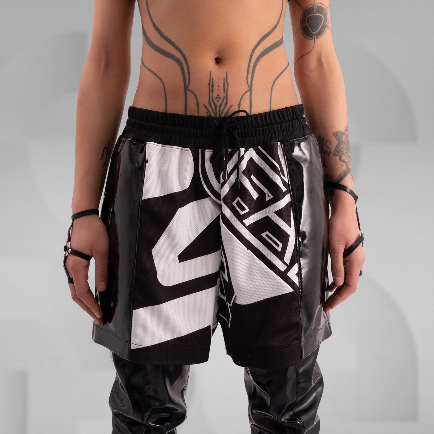 Close-up of a model wearing black and white graphic shorts with a bold geometric design, paired with glossy black PVC side panels. Styled with statement rings, bracelets, and intricate body tattoos, creating an edgy, futuristic streetwear look.