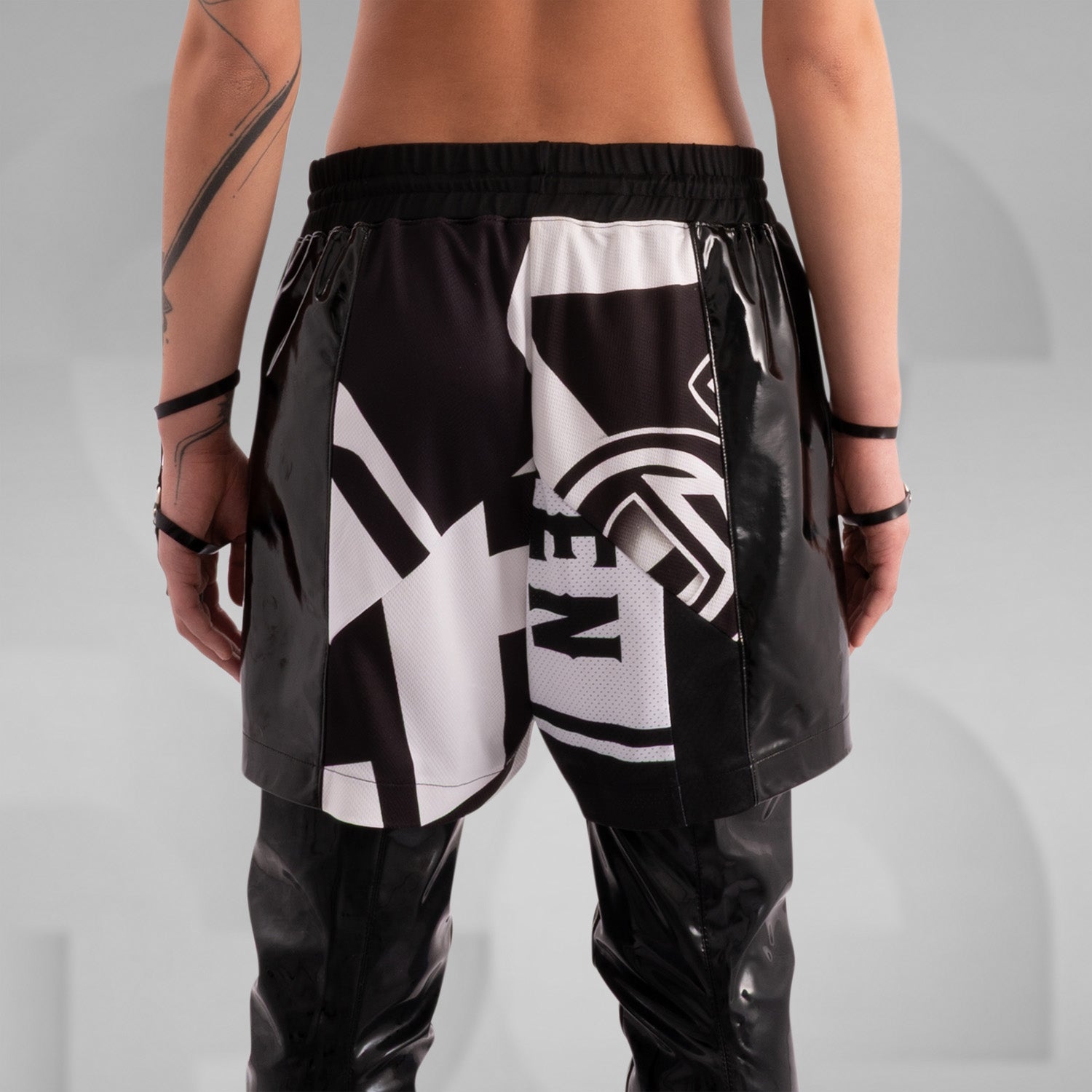 Close-up of a model wearing black and white graphic shorts with a bold geometric design, paired with glossy black PVC side panels. Styled with statement rings, bracelets, and intricate body tattoos, creating an edgy, futuristic streetwear look.