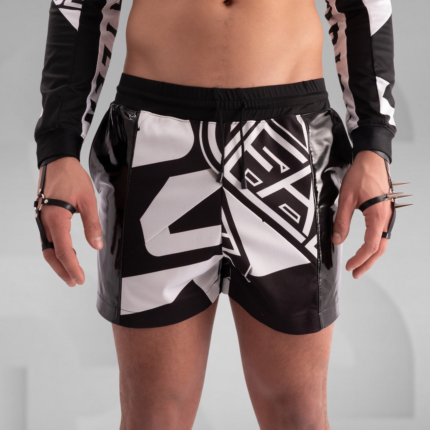 Close-up of a model wearing black and white graphic shorts with a bold geometric design, paired with glossy black PVC side panels. Styled with statement rings, bracelets, and intricate body tattoos, creating an edgy, futuristic streetwear look.