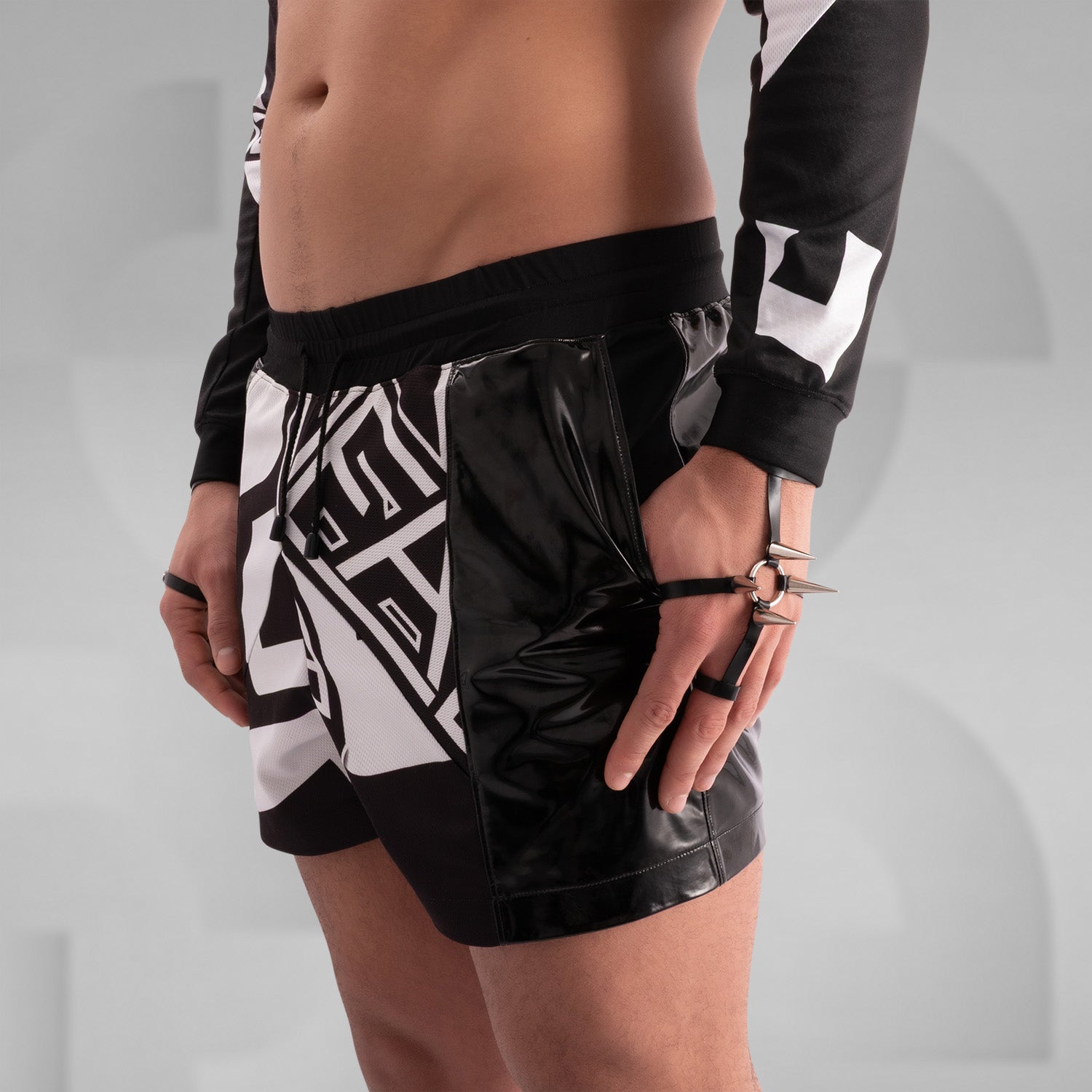 Close-up of a model wearing black and white graphic shorts with a bold geometric design, paired with glossy black PVC side panels. Styled with statement rings, bracelets, and intricate body tattoos, creating an edgy, futuristic streetwear look.