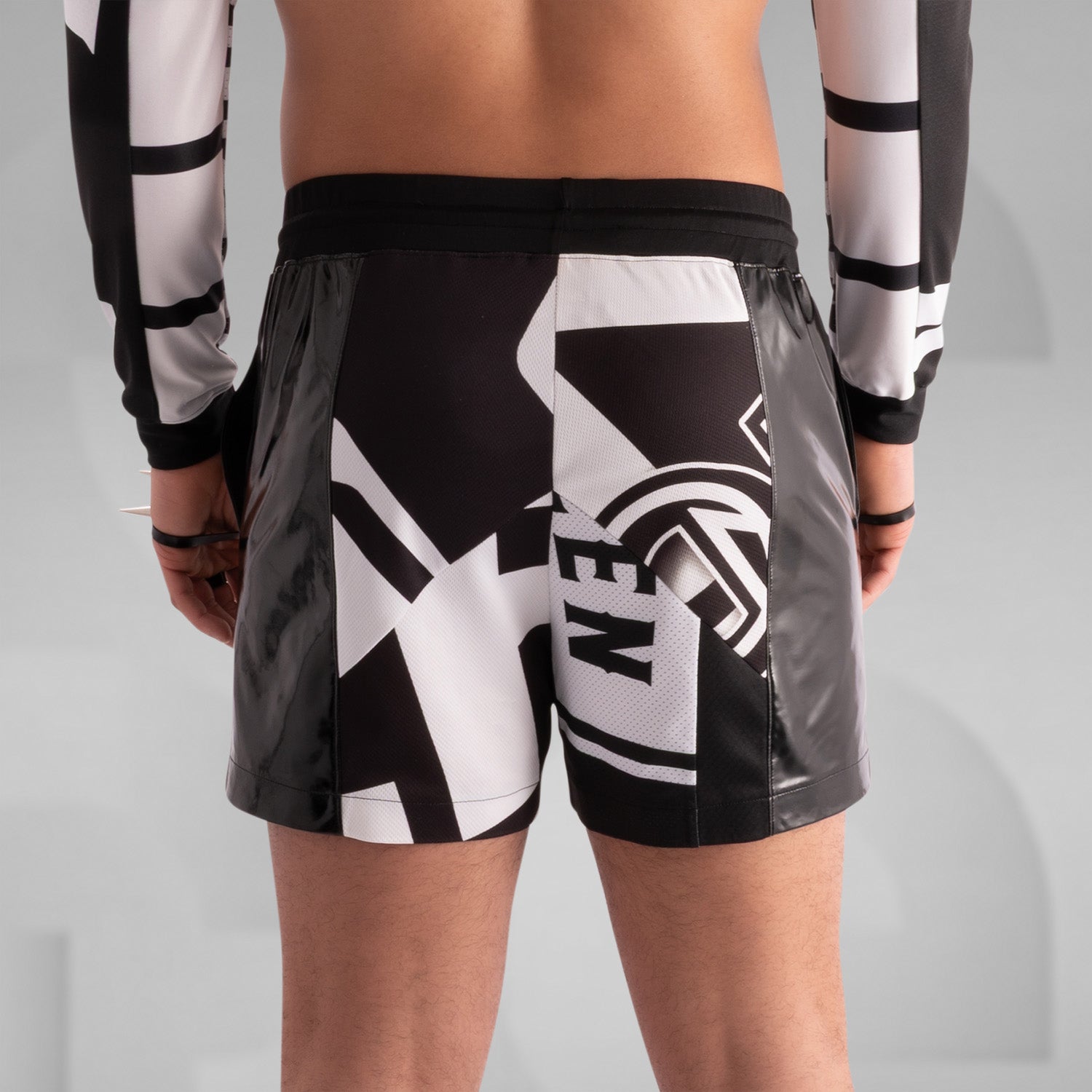 Close-up of a model wearing black and white graphic shorts with a bold geometric design, paired with glossy black PVC side panels. Styled with statement rings, bracelets, and intricate body tattoos, creating an edgy, futuristic streetwear look.