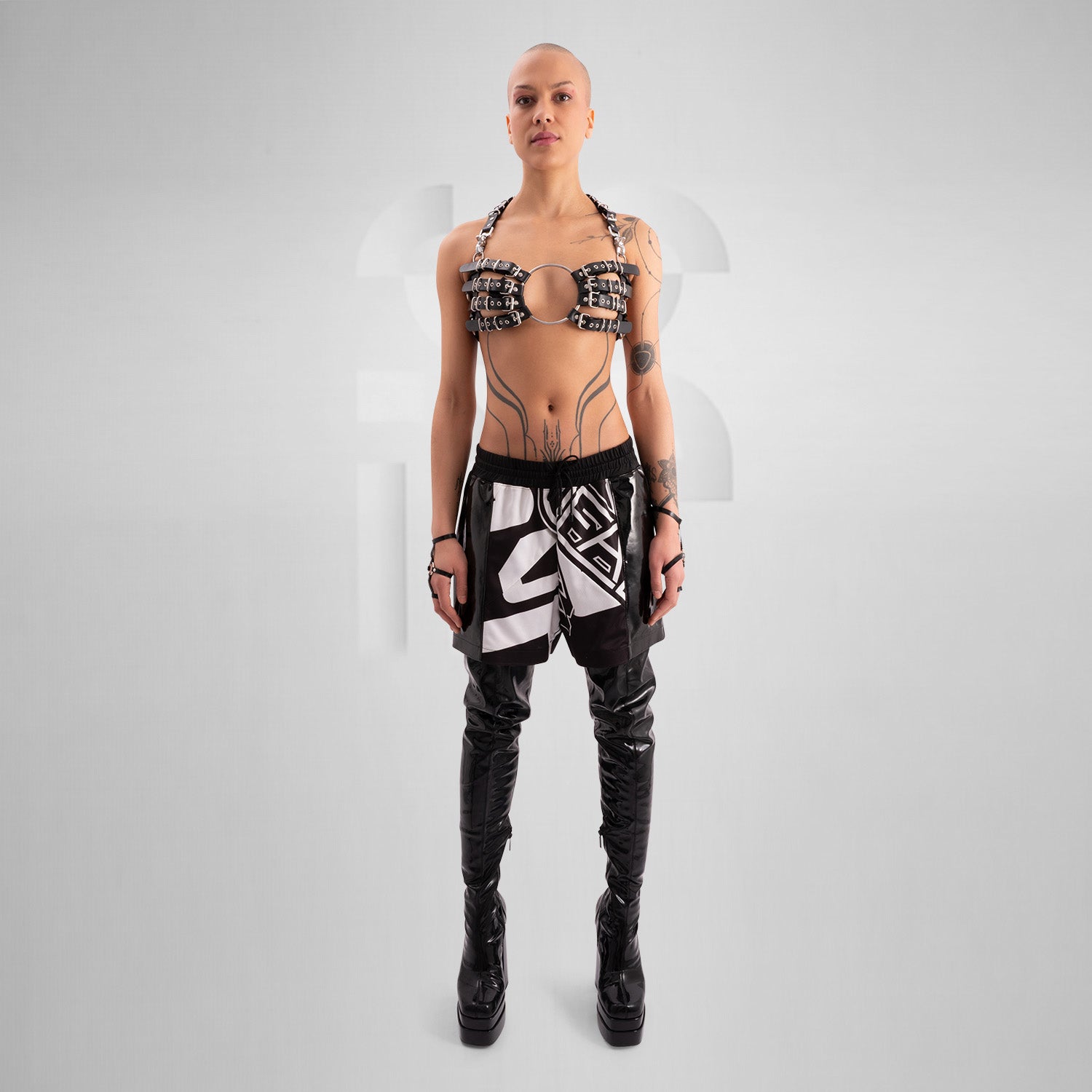 This image features a person wearing a unique and edgy outfit. The top is minimal and metallic, resembling a harness, while the skirt has bold black and white geometric designs. The outfit is completed with tall, shiny black thigh-high boots. The styling appears to be avant-garde and fashion-forward.