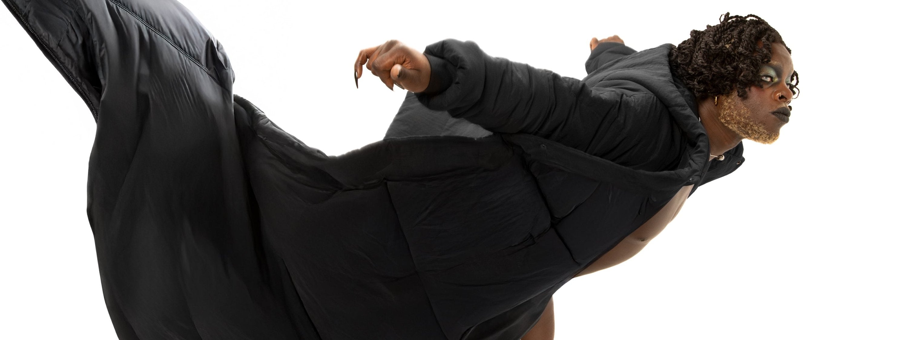 A in a dramatic, flowing black puffer coat, captured in motion as if it's billowing around them. The outfit contrasts with their bold makeup and styled hair. The background is pure white, highlighting the striking silhouette and the fluidity of the coat, adding a sense of dynamic movement to the image.