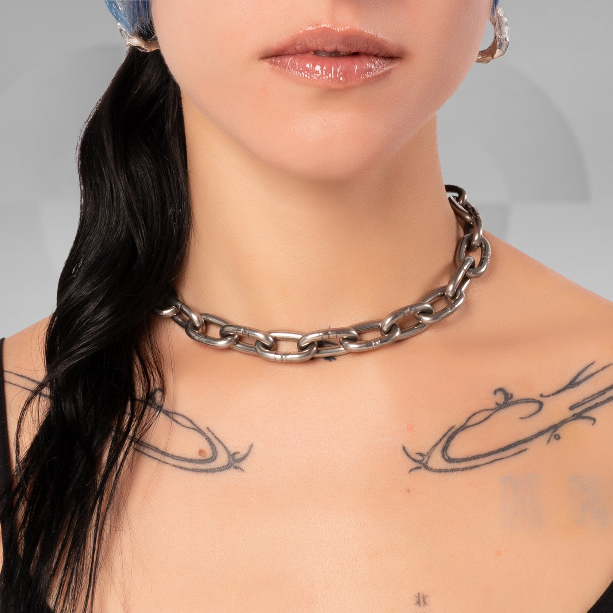 A close-up of a model wearing a chunky silver-toned metal chain choker necklace. The model has black hair in a low ponytail, visible skin with black line-art tattoos on the chest, and is wearing silver hoop earrings.