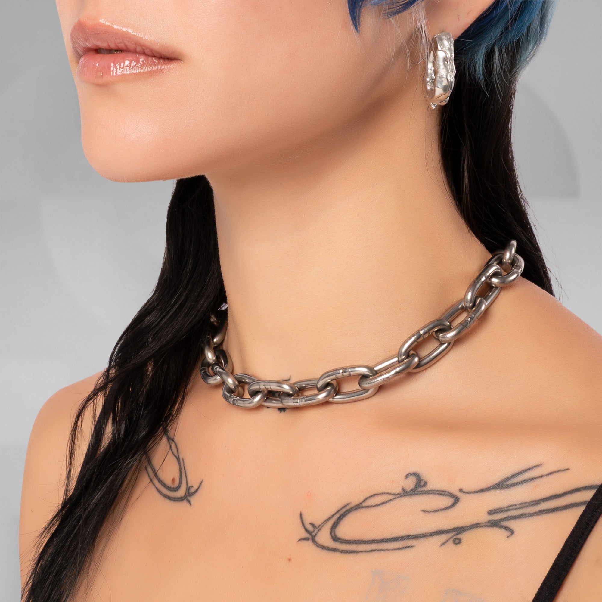 A close-up of a model wearing a chunky silver chain choker necklace, with black hair, blue highlights, and visible chest tattoos against a soft gray background.