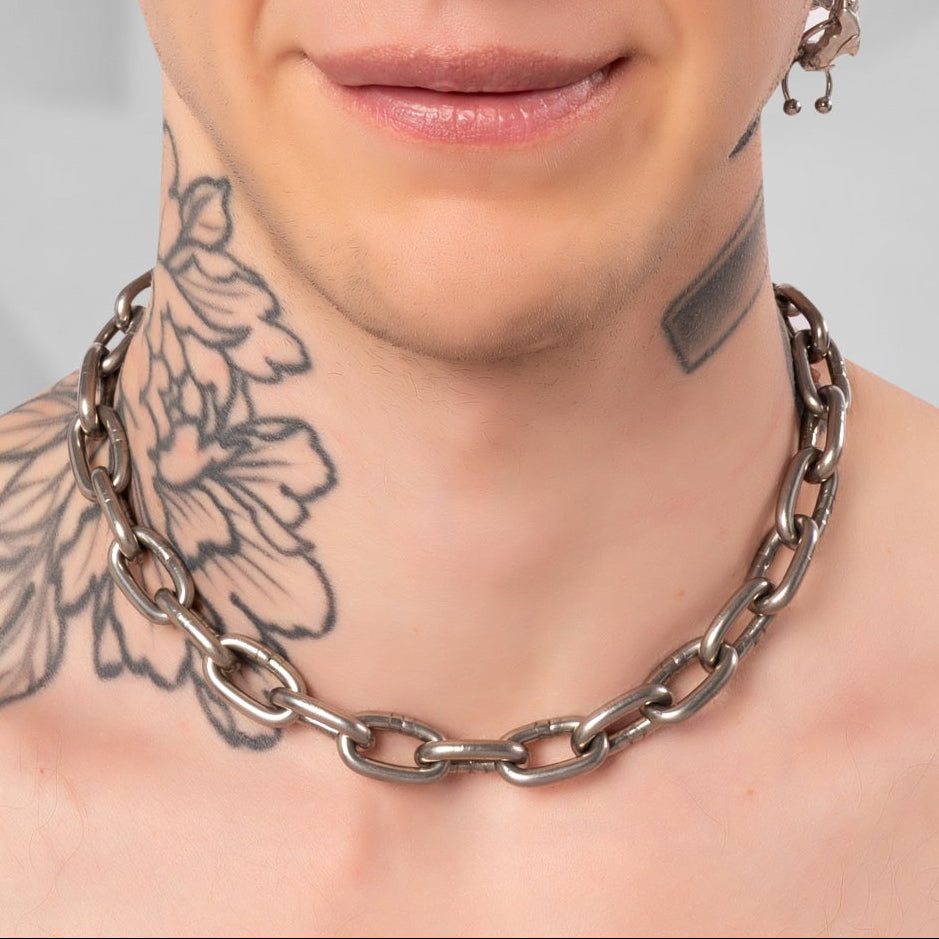 A close-up portrait of a man with a buzz cut, wearing a chunky silver chain necklace and multiple silver earrings, set against a gray background with a subtle circular graphic.