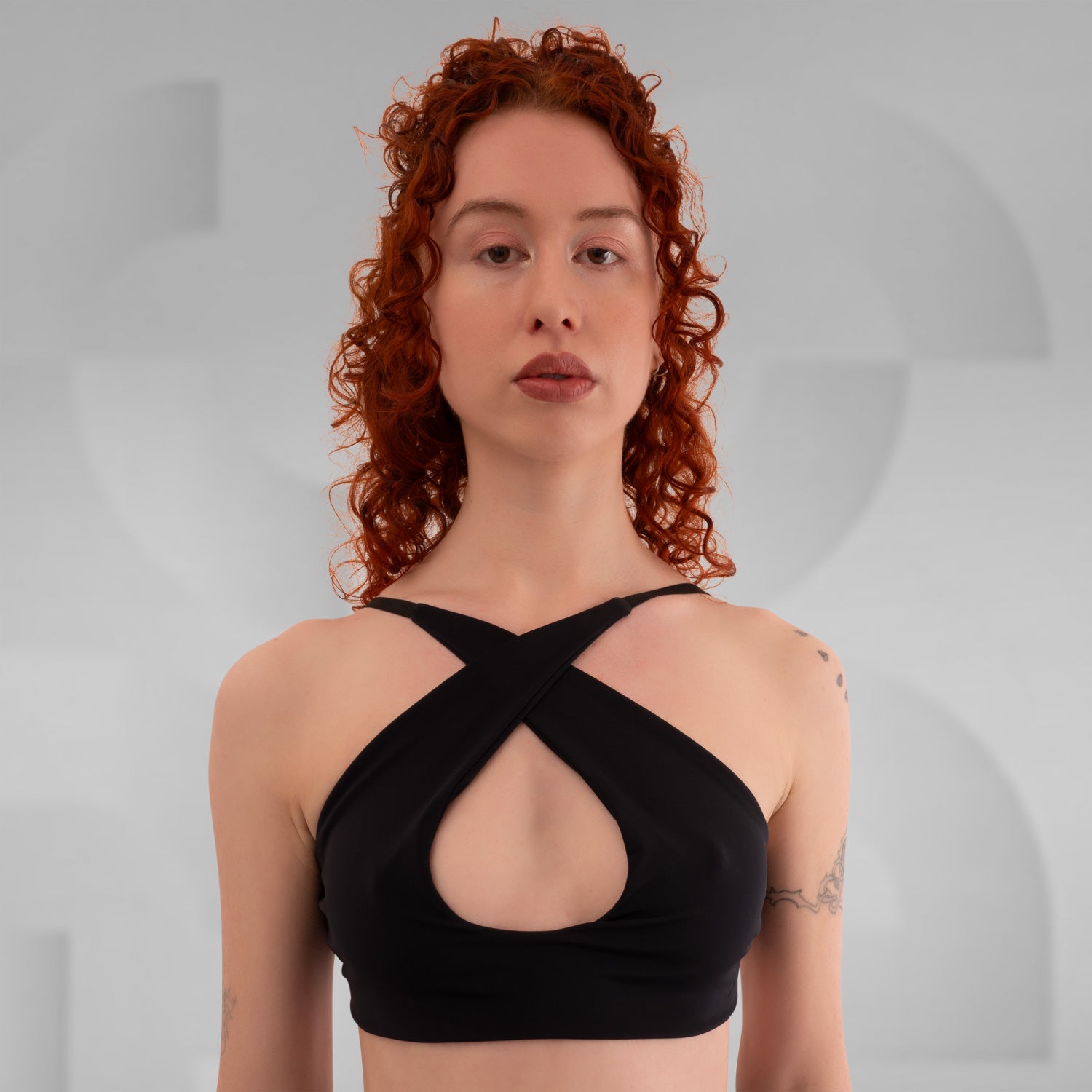 This image depicts a young woman with curly red hair wearing a black crisscross halter-style top that features a cutout detail at the chest. Her expression is neutral, and she stands against a minimalist gray background with soft, abstract geometric patterns. Visible tattoos on her upper arm add subtle detail to her appearance. The composition of the image is focused on the subject's upper body, emphasizing the stylish and modern design of her top.
