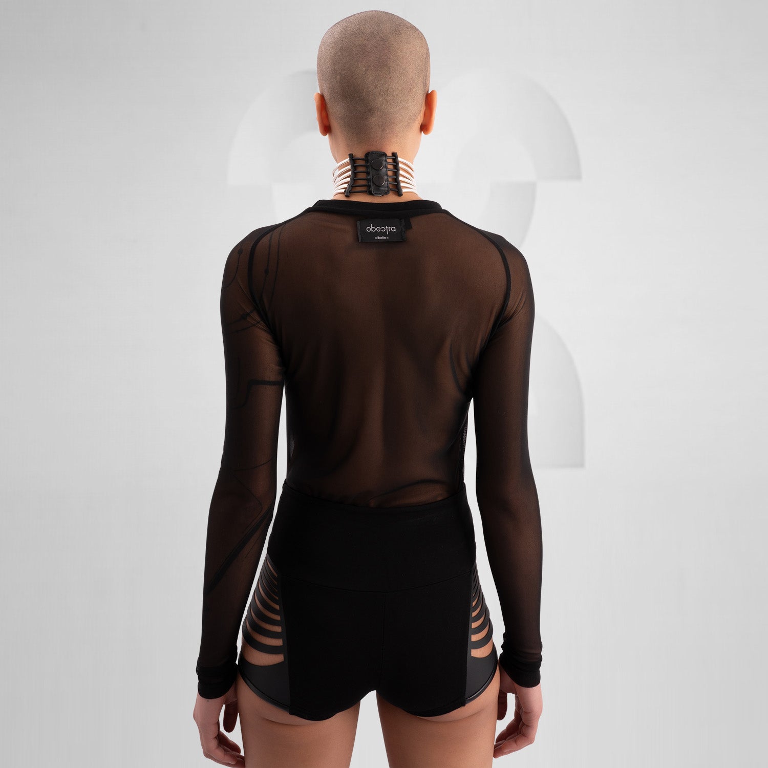 Model wearing a bold, futuristic black sheer long-sleeve top with a high neckline, paired with black cut-out shorts. The look is completed with a statement metallic choker and nipple pasties, creating an edgy, avant-garde aesthetic. Perfect for techno, festival, and alternative fashion