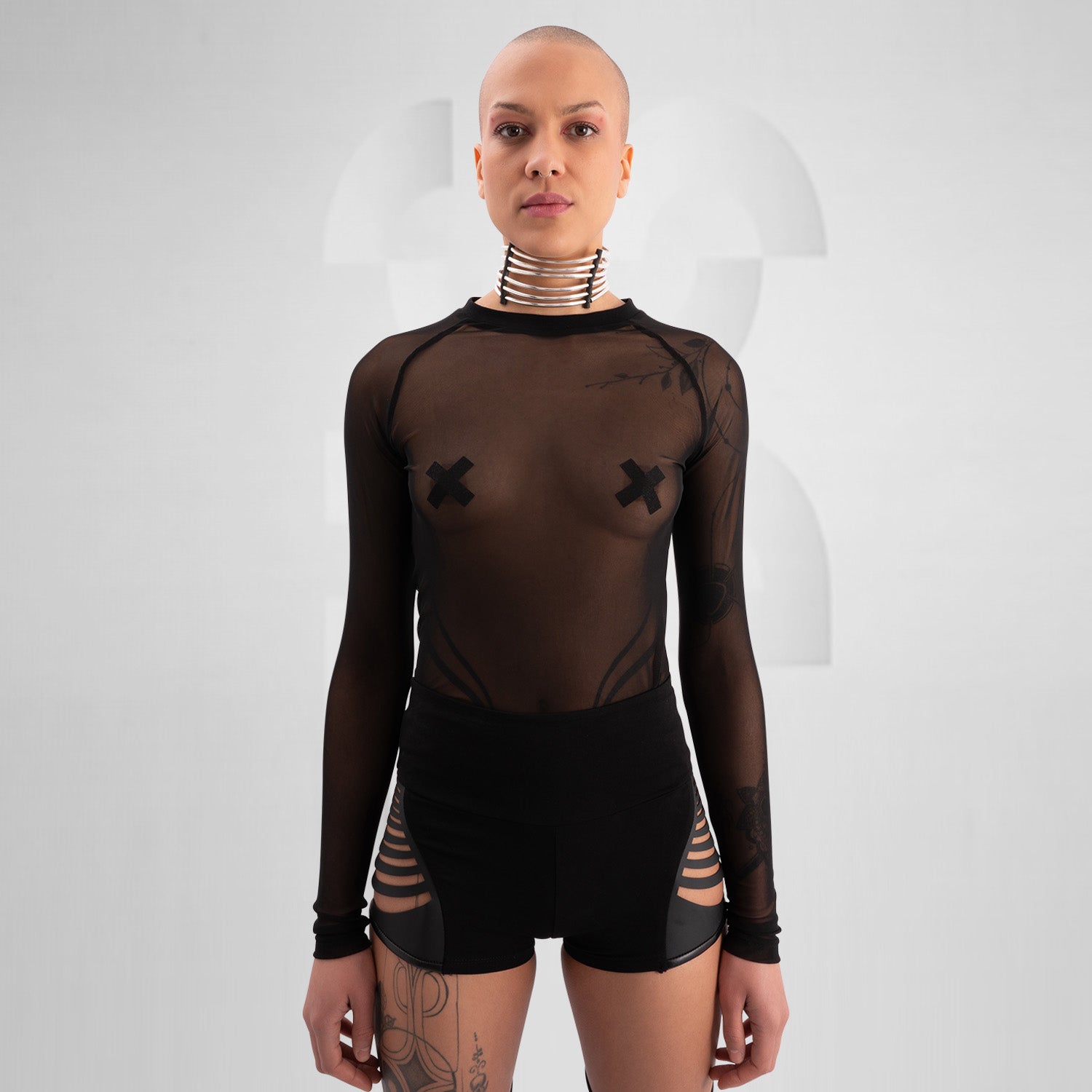 Model wearing a bold, futuristic black sheer long-sleeve top with a high neckline, paired with black cut-out shorts. The look is completed with a statement metallic choker and nipple pasties, creating an edgy, avant-garde aesthetic. Perfect for techno, festival, and alternative fashion