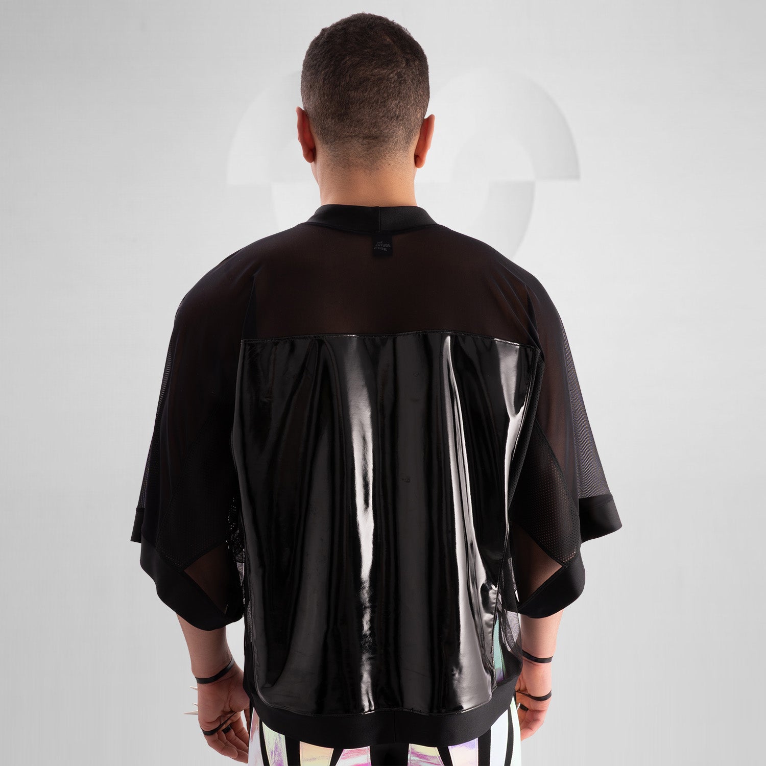 The image features a person wearing a striking outfit that combines a black, glossy top with sheer mesh sleeves, creating a bold and modern aesthetic. The top's shiny material adds an eye-catching element, while the coordinating shorts feature angular holographic designs that reflect light beautifully. This look embodies a dynamic blend of avant-garde fashion and street style, perfect for making a statement.