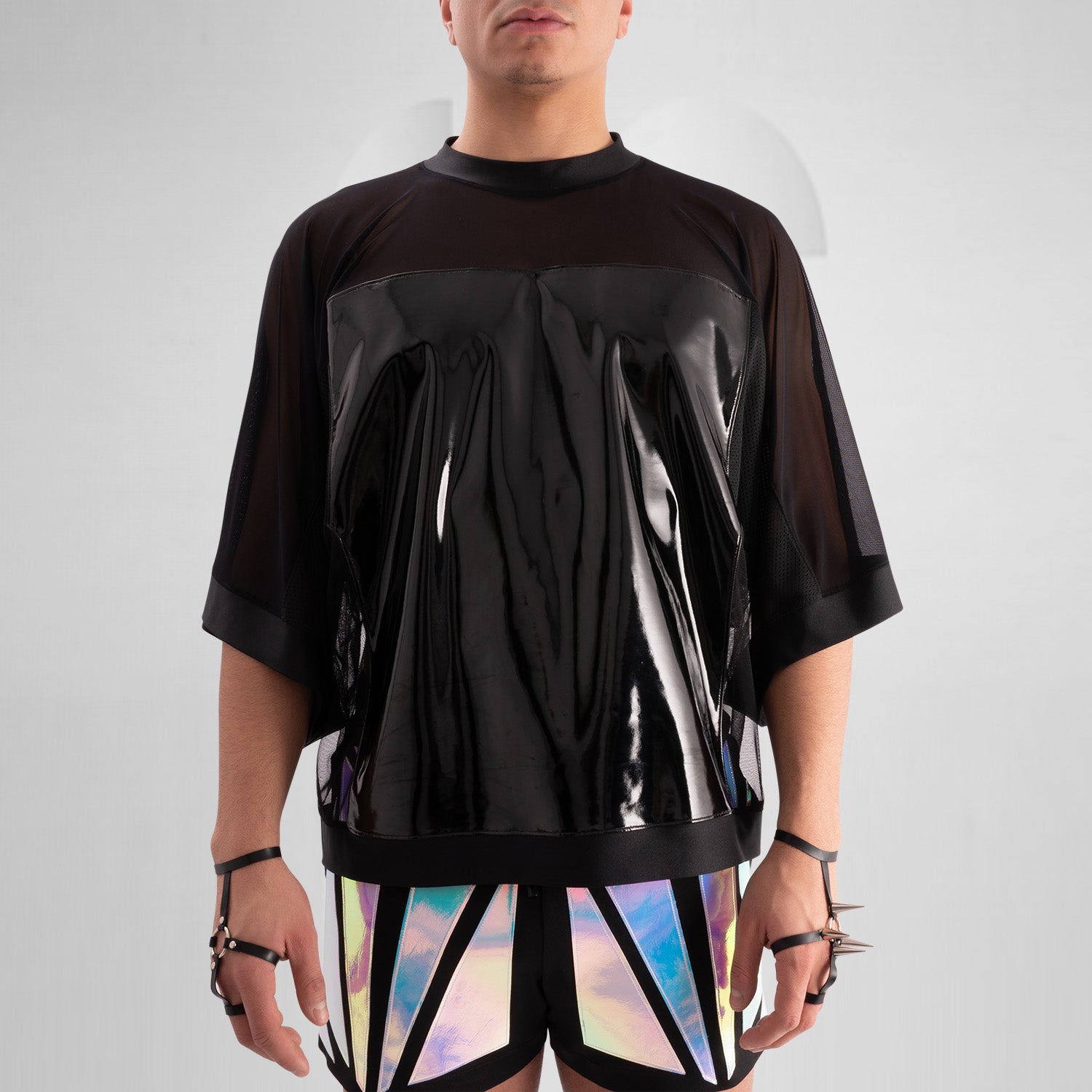 The image features a person wearing a striking outfit that combines a black, glossy top with sheer mesh sleeves, creating a bold and modern aesthetic. The top's shiny material adds an eye-catching element, while the coordinating shorts feature angular holographic designs that reflect light beautifully. This look embodies a dynamic blend of avant-garde fashion and street style, perfect for making a statement.