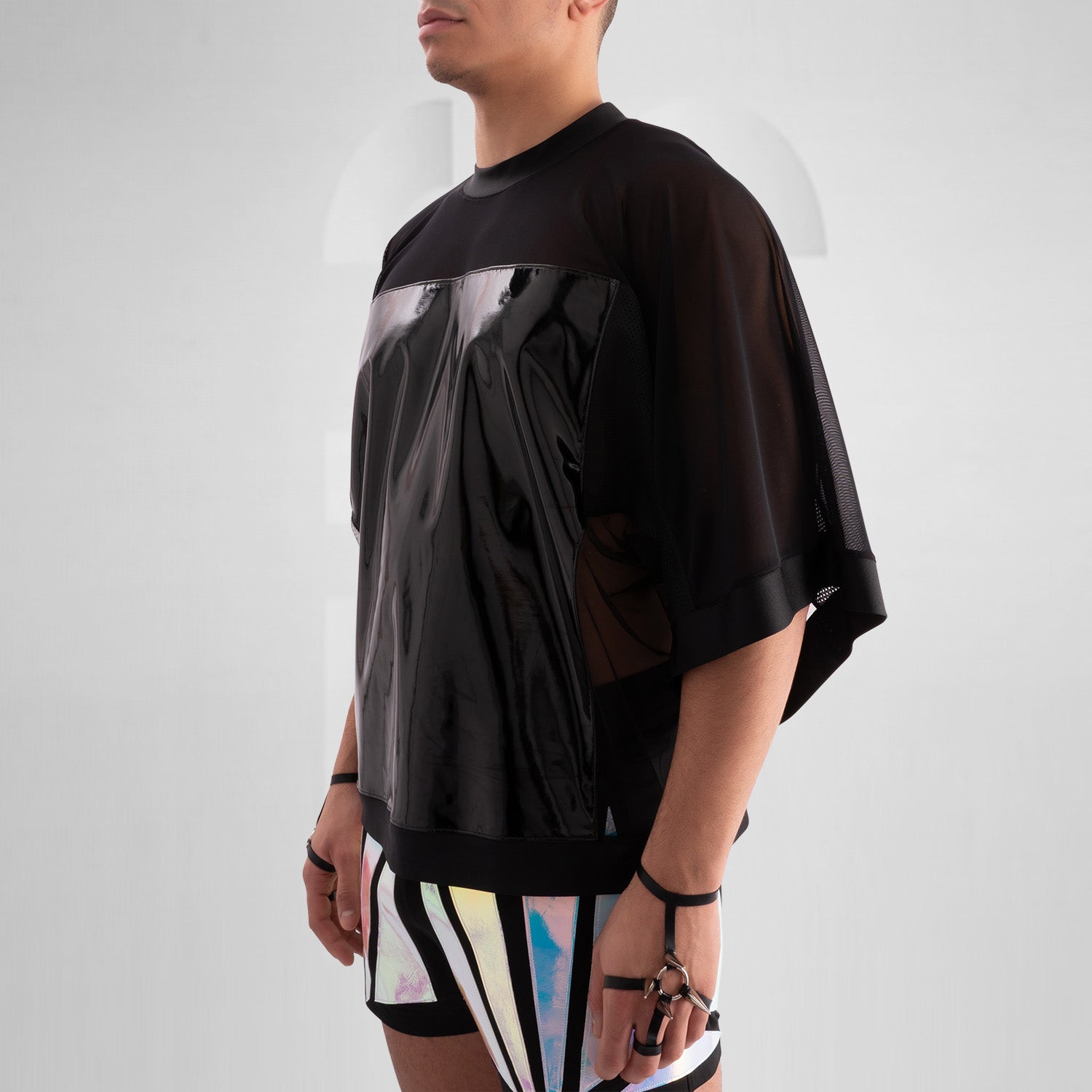 The image features a person wearing a striking outfit that combines a black, glossy top with sheer mesh sleeves, creating a bold and modern aesthetic. The top's shiny material adds an eye-catching element, while the coordinating shorts feature angular holographic designs that reflect light beautifully. This look embodies a dynamic blend of avant-garde fashion and street style, perfect for making a statement.