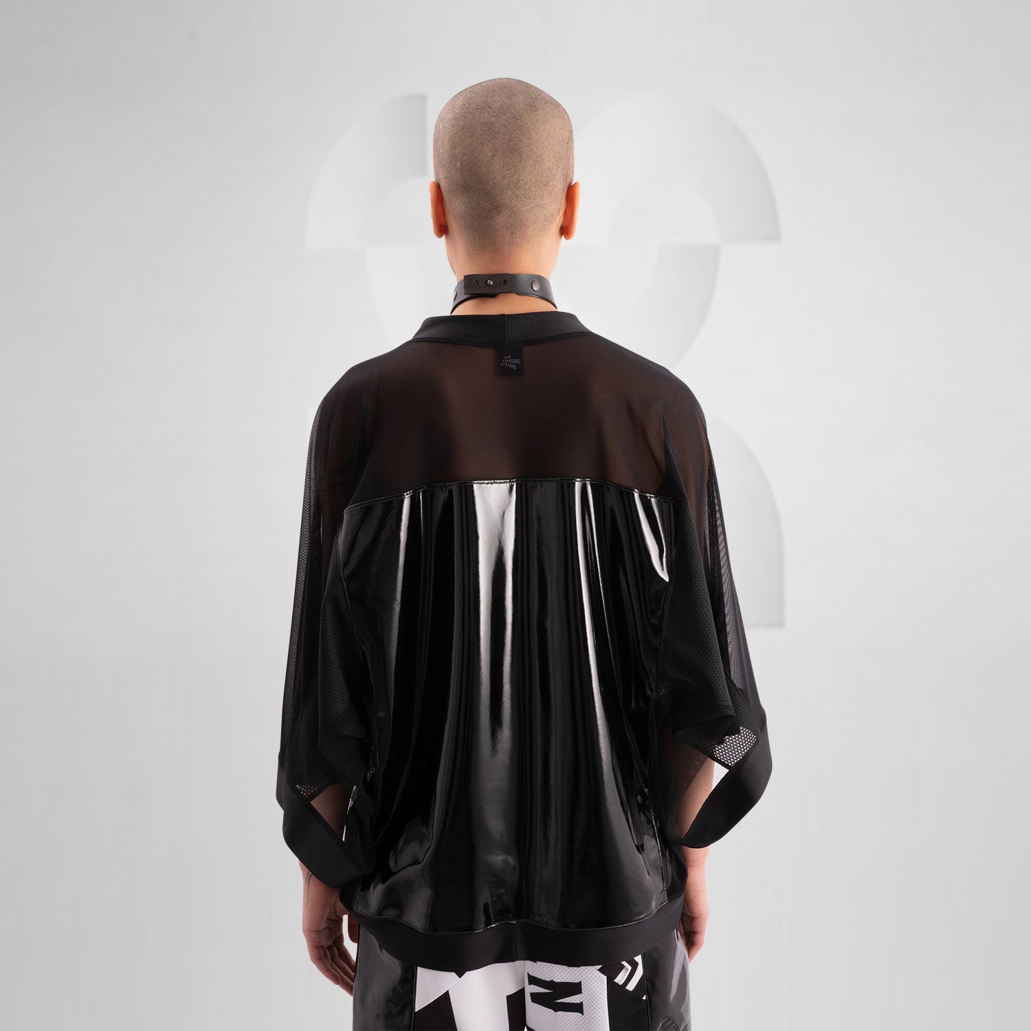 The image features a person wearing a striking outfit that combines a black, glossy top with sheer mesh sleeves, creating a bold and modern aesthetic. The top's shiny material adds an eye-catching element, while the coordinating shorts feature angular holographic designs that reflect light beautifully. This look embodies a dynamic blend of avant-garde fashion and street style, perfect for making a statement.