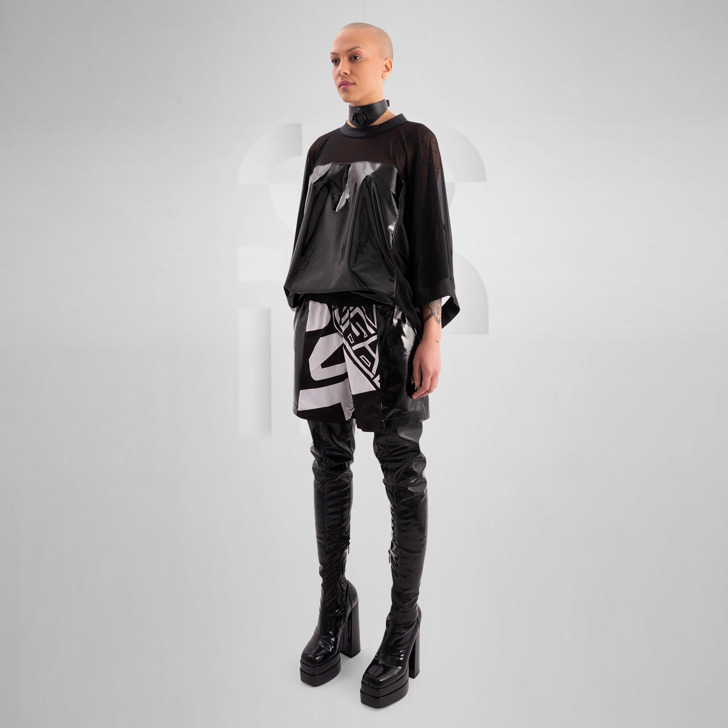 The image features a person wearing a striking outfit that combines a black, glossy top with sheer mesh sleeves, creating a bold and modern aesthetic. The top's shiny material adds an eye-catching element, while the coordinating shorts feature angular holographic designs that reflect light beautifully. This look embodies a dynamic blend of avant-garde fashion and street style, perfect for making a statement.