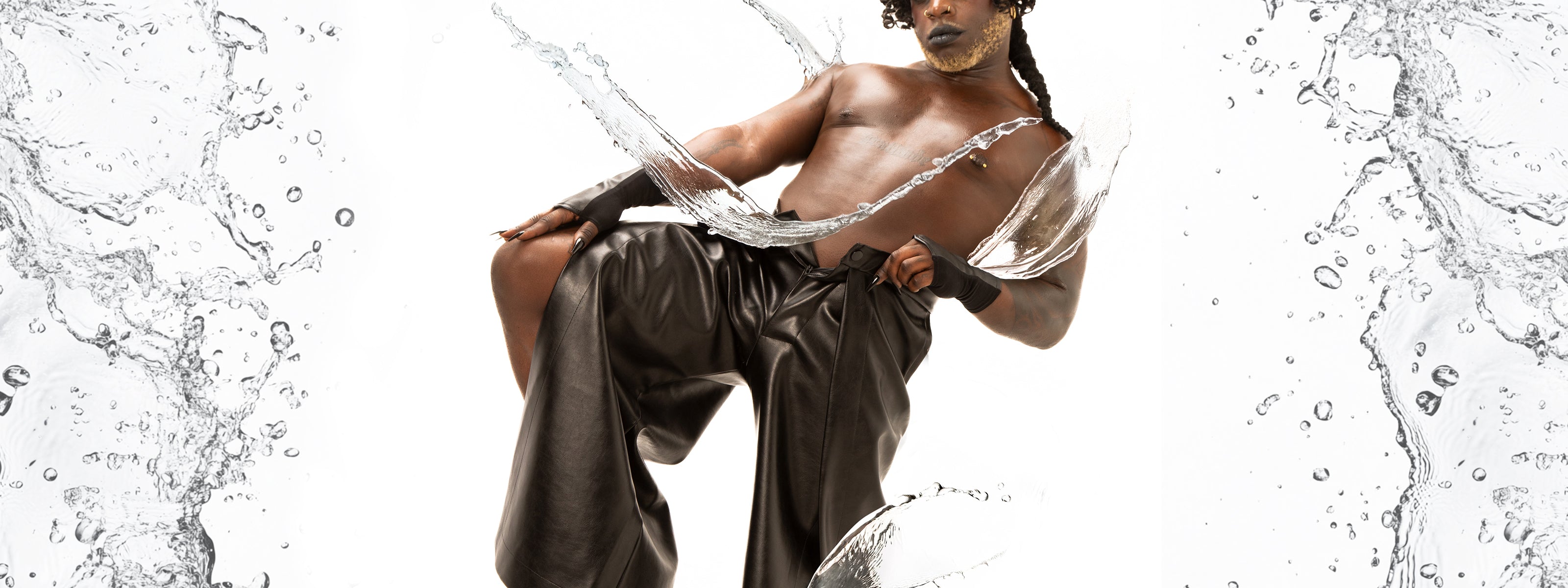 posed, showcasing a bare chest paired with flowing, dark leather pants. The model’s hands are adorned with stylish accessories, and their hair is styled dramatically. Surrounding the figure are dynamic splashes of water that create a sense of movement, set against a clean white background that highlights the boldness of the outfit and the model's presence.