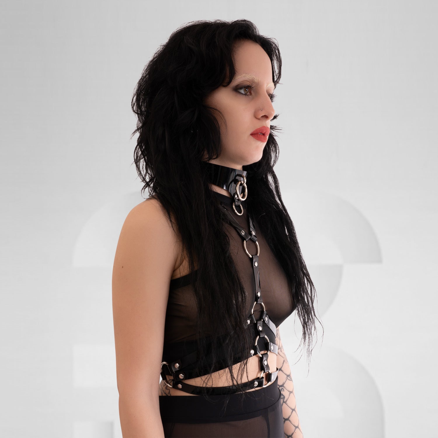 This image showcases a model wearing a striking black harness top that features a combination of sleek straps and bold detailing. The design includes eye-catching golden accents that enhance its edgy aesthetic, perfectly blending style and attitude. Crafted from high-quality materials, the harness contours the body beautifully while ensuring comfort and movement.