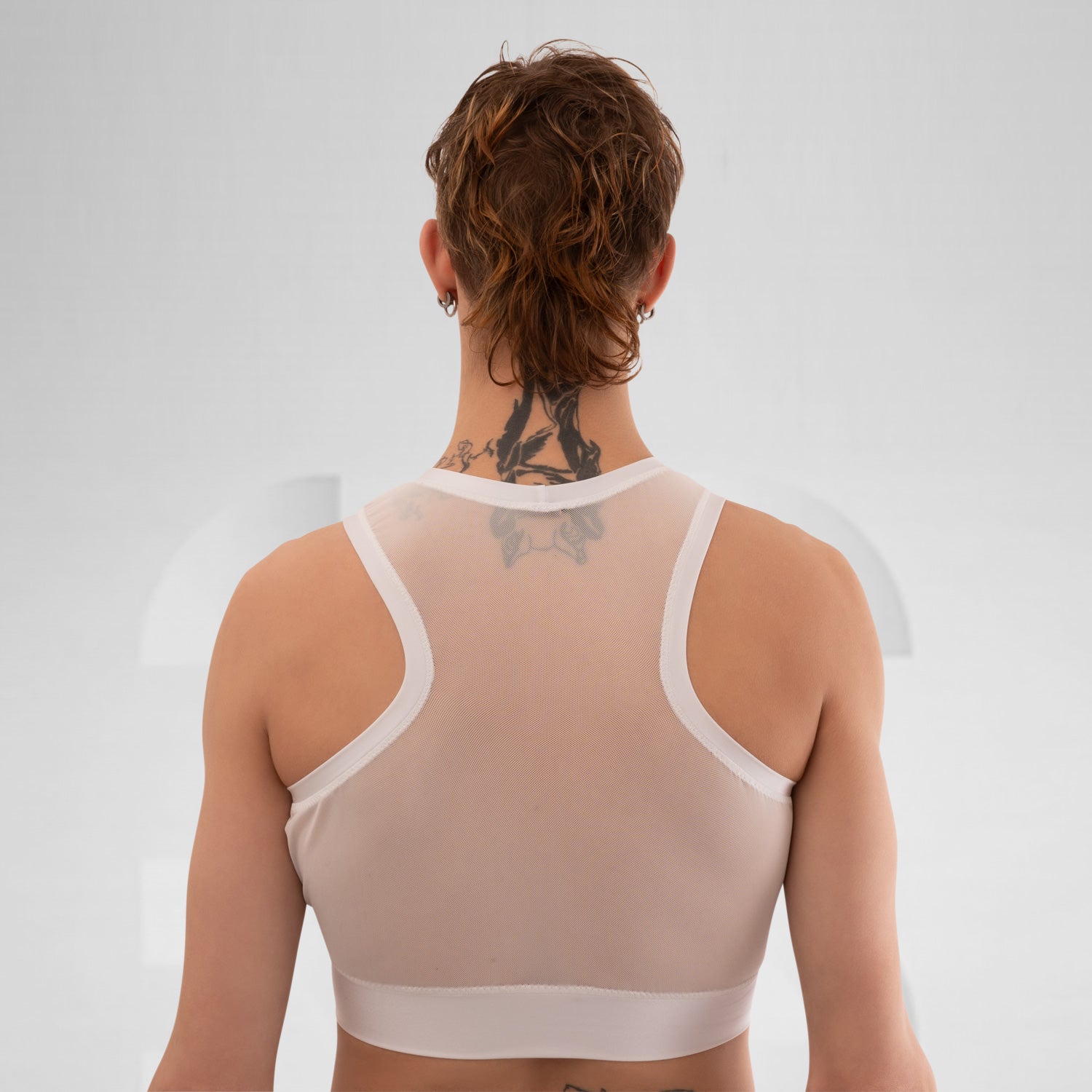 The side profile of a model wearing a translucent white top with a modern, sporty design. The top is complemented by a choker that has a gold detail, adding an edgy touch. The model has short, vibrant red hair and dramatic makeup, enhancing their bold look. The neutral background focuses attention on the outfit and the striking features of the model.