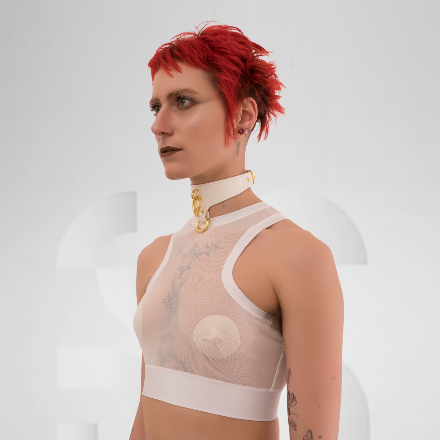 The side profile of a model wearing a translucent white top with a modern, sporty design. The top is complemented by a choker that has a gold detail, adding an edgy touch. The model has short, vibrant red hair and dramatic makeup, enhancing their bold look. The neutral background focuses attention on the outfit and the striking features of the model.