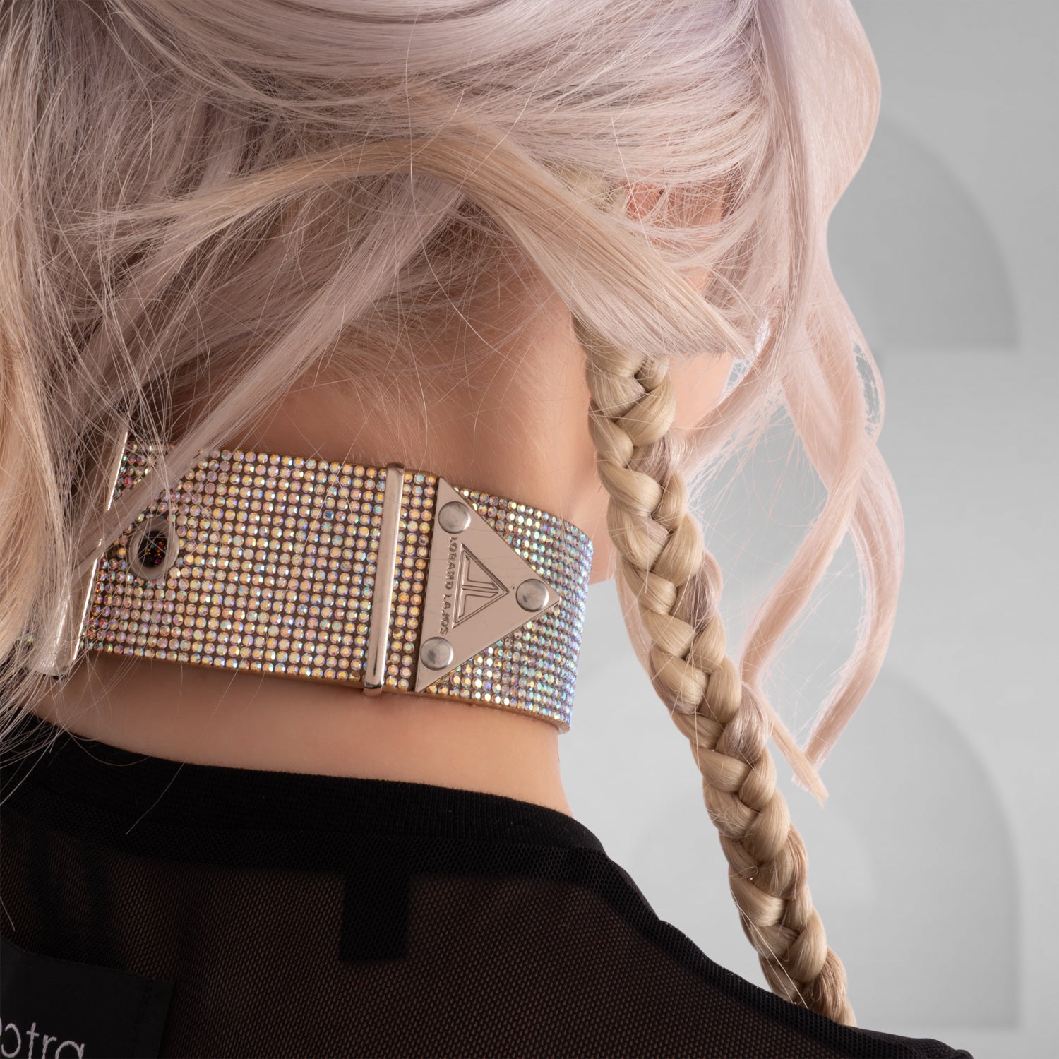 This close-up image highlights a woman with a daring, edgy style. She has platinum blonde hair styled in loose waves with a braid on one side. She's wearing a sparkling, multi-colored rhinestone choker around her neck, which adds a glamorous, bold statement to her look. Her makeup features vibrant red lipstick matching her accessories, and she has multiple facial piercings, including a septum ring and dermal piercings below her lips, emphasizing her alternative, fashion-forward aesthetic.