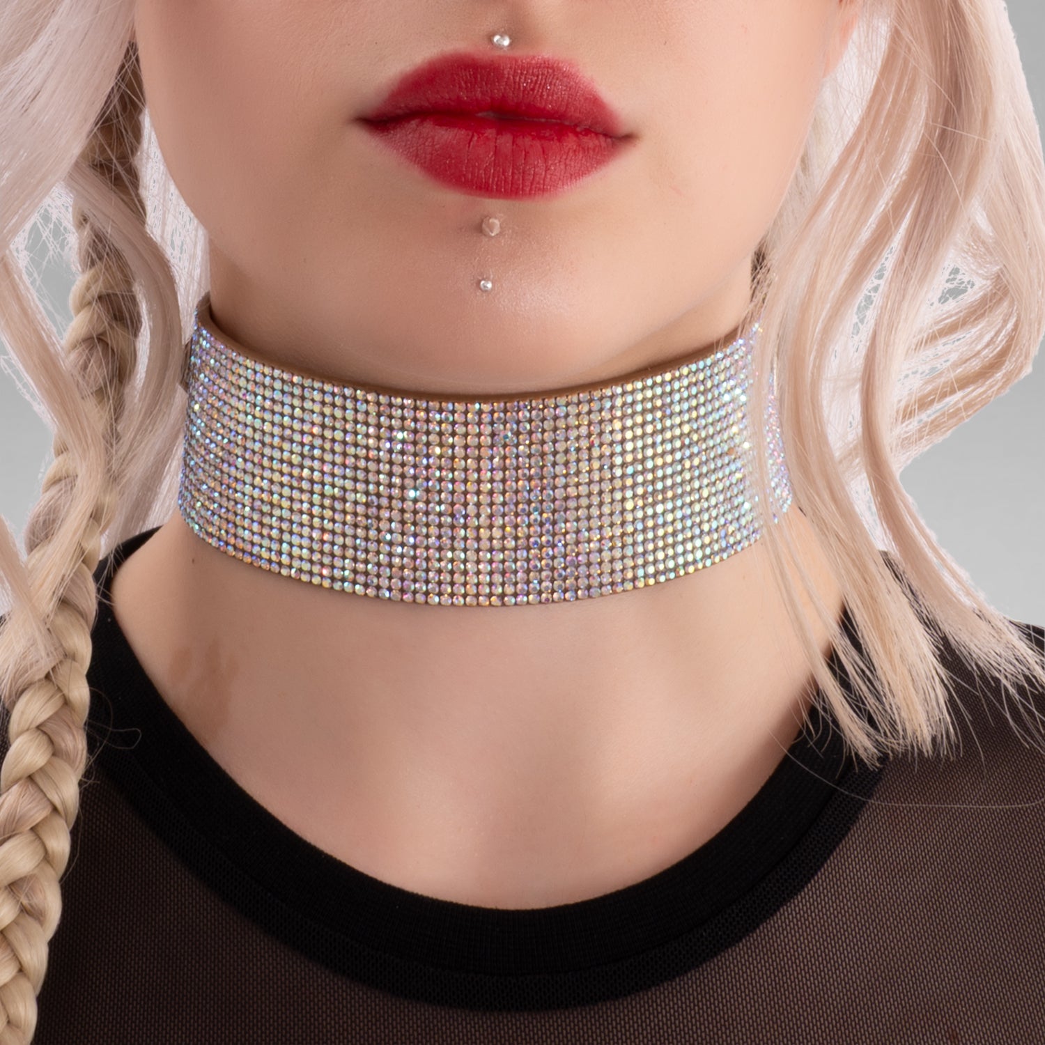 This close-up image highlights a woman with a daring, edgy style. She has platinum blonde hair styled in loose waves with a braid on one side. She's wearing a sparkling, multi-colored rhinestone choker around her neck, which adds a glamorous, bold statement to her look. Her makeup features vibrant red lipstick matching her accessories, and she has multiple facial piercings, including a septum ring and dermal piercings below her lips, emphasizing her alternative, fashion-forward aesthetic.