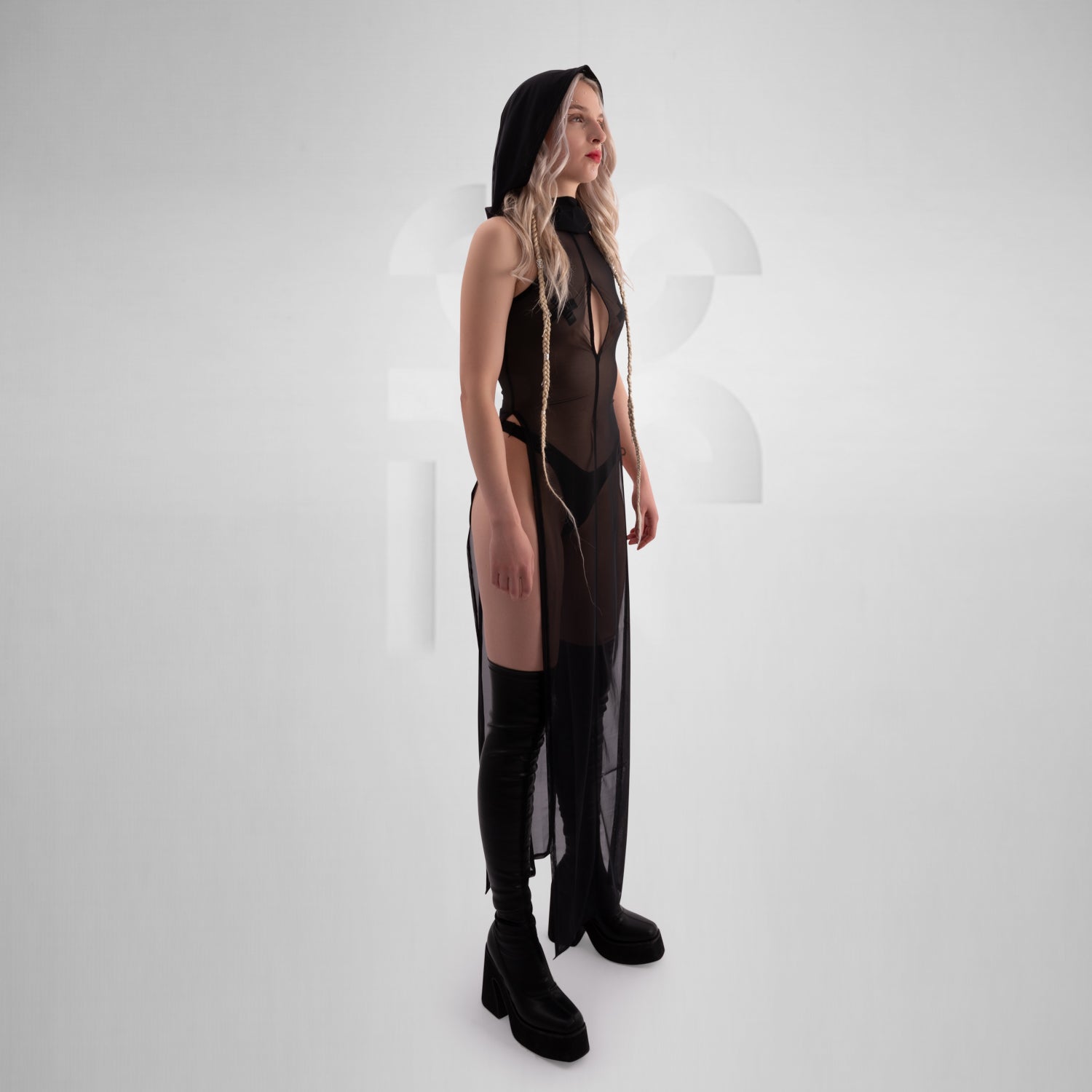 Model wearing a sheer black hooded dress with keyhole chest detail, belted at the waist with a chunky chain-accented rubber belt, paired with black platform boots and visible black lingerie — edgy, gothic-inspired look perfect for clubwear or alternative fashion.
