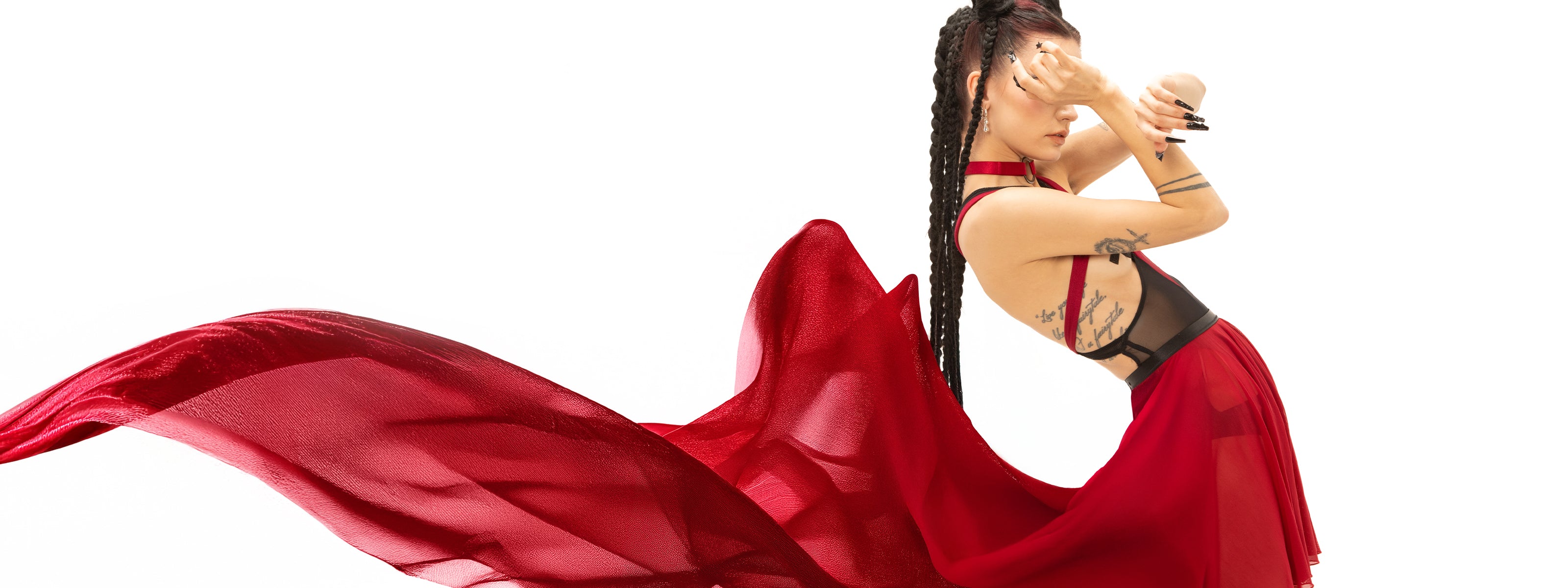 pose featuring a model dressed in a sheer red flowy dress with intricate layering. The model's hair is styled in braids, and they are partially obscuring their face with a hand, adding an element of intrigue. The vibrant fabric flows around them, creating a sense of movement against the clean white background, highlighting their bold look and artistic expression.