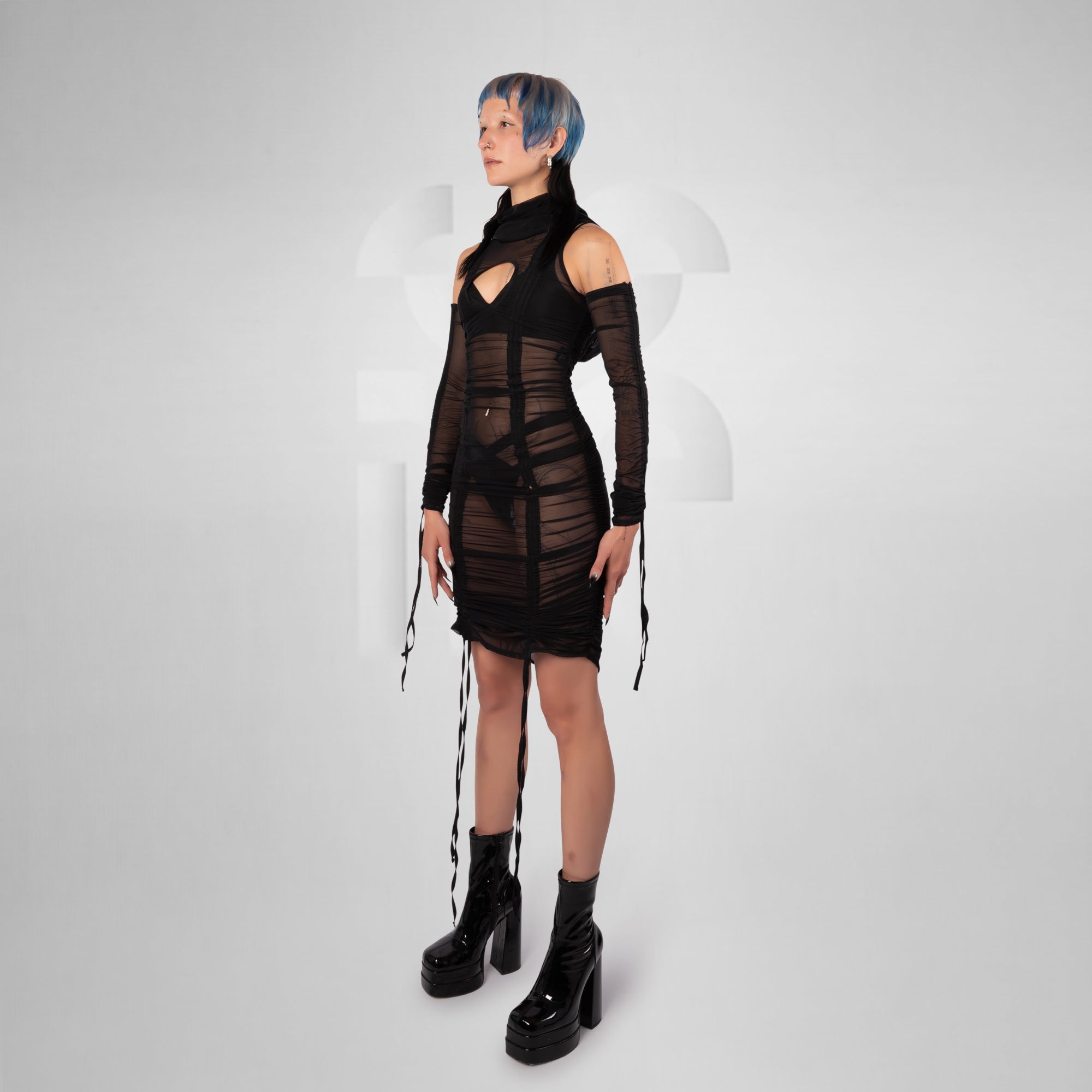 A model with short blue hair stands in a studio wearing a black sheer mesh mini dress with cutouts, long sleeves, and dangling tie details, paired with black platform ankle boots, against a light gray background.