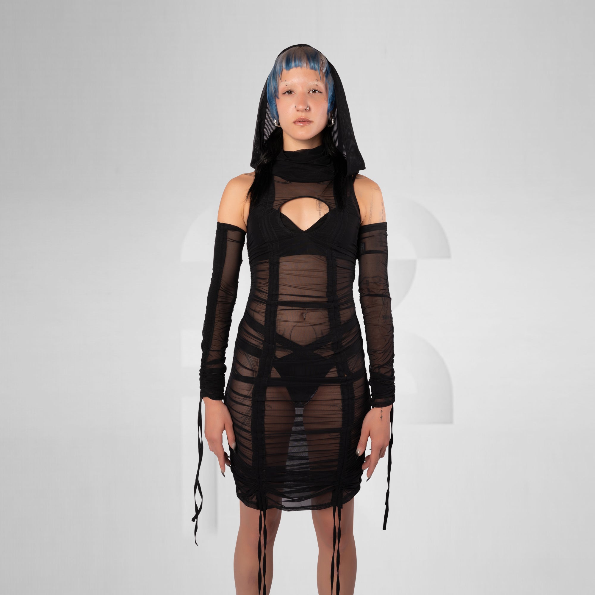 A model with blue-tinted hair wearing a black sheer mesh mini dress with long sleeves, a high neck, and front cutouts, standing against a light gray studio background.
