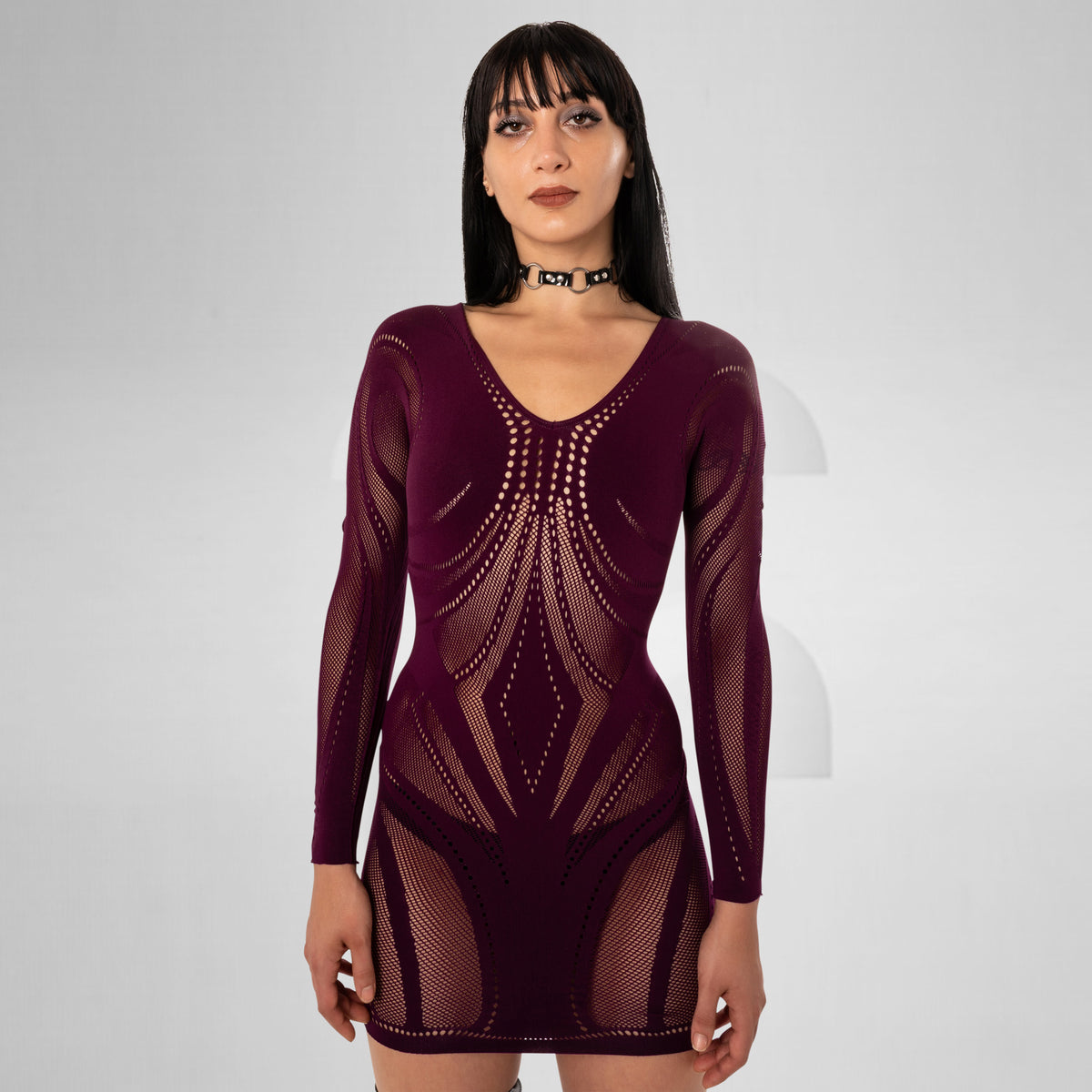 A woman modeling a long-sleeve, bodycon mini dress made of sheer burgundy mesh with geometric cutout patterns, set against a plain light gray studio background.