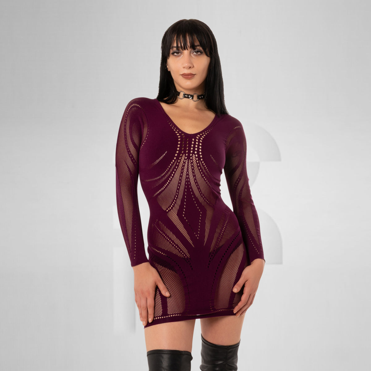A woman modeling a long-sleeve, sheer mesh mini dress in deep burgundy with geometric cutout patterns, paired with black knee-high boots and a black choker against a light gray studio background.