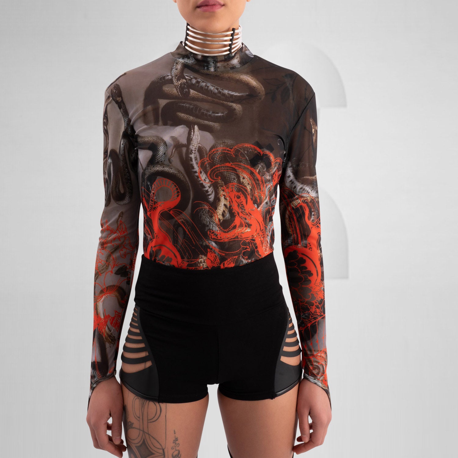 The image showcases a striking outfit featuring a long-sleeve top with an eye-catching design of swirling octopuses in dark hues, accented with vibrant red. The top is complemented by unique metallic neck detailing that adds an edgy flair. Paired with black high-waisted shorts featuring cut-out details, this look exudes confidence and individuality. Ideal for making a statement on the dancefloor or at a fashion-forward event, the ensemble beautifully blends art and style for a captivating appearance.