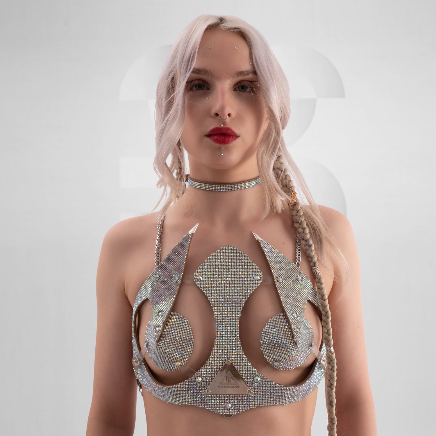 This image features a woman with platinum blonde hair styled in loose waves and a single braid on one side. She is wearing a highly decorative and futuristic top made of shimmering, rhinestone-encrusted materials that cover her chest and shoulders, with pointed embellishments extending outward. Her makeup includes bold red lipstick, and she has multiple facial piercings, including a septum ring and dermal piercings near her eyes and lips. 