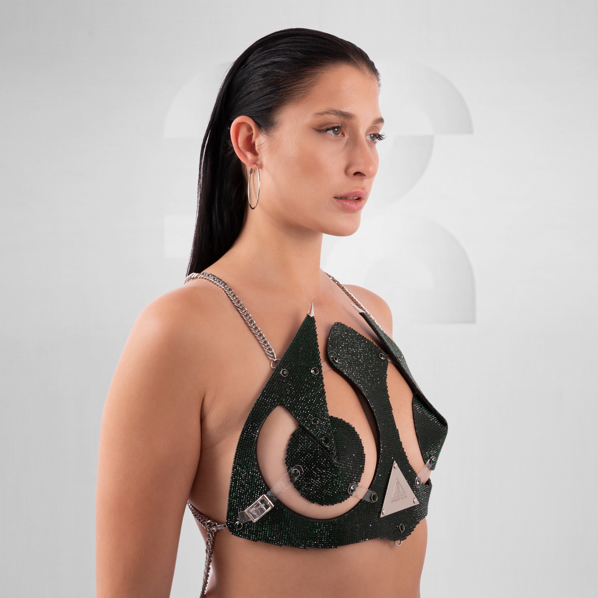 Woman wearing an avant-garde red crystal bra top with geometric cutouts and silver chain details, styled for festivals or high-fashion clubwear; the top features bold, futuristic design elements with a metallic triangle logo centerpiece.