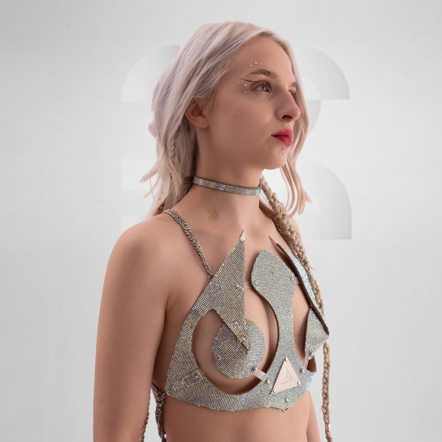 This image features a woman with platinum blonde hair styled in loose waves and a single braid on one side. She is wearing a highly decorative and futuristic top made of shimmering, rhinestone-encrusted materials that cover her chest and shoulders, with pointed embellishments extending outward. Her makeup includes bold red lipstick, and she has multiple facial piercings, including a septum ring and dermal piercings near her eyes and lips. 