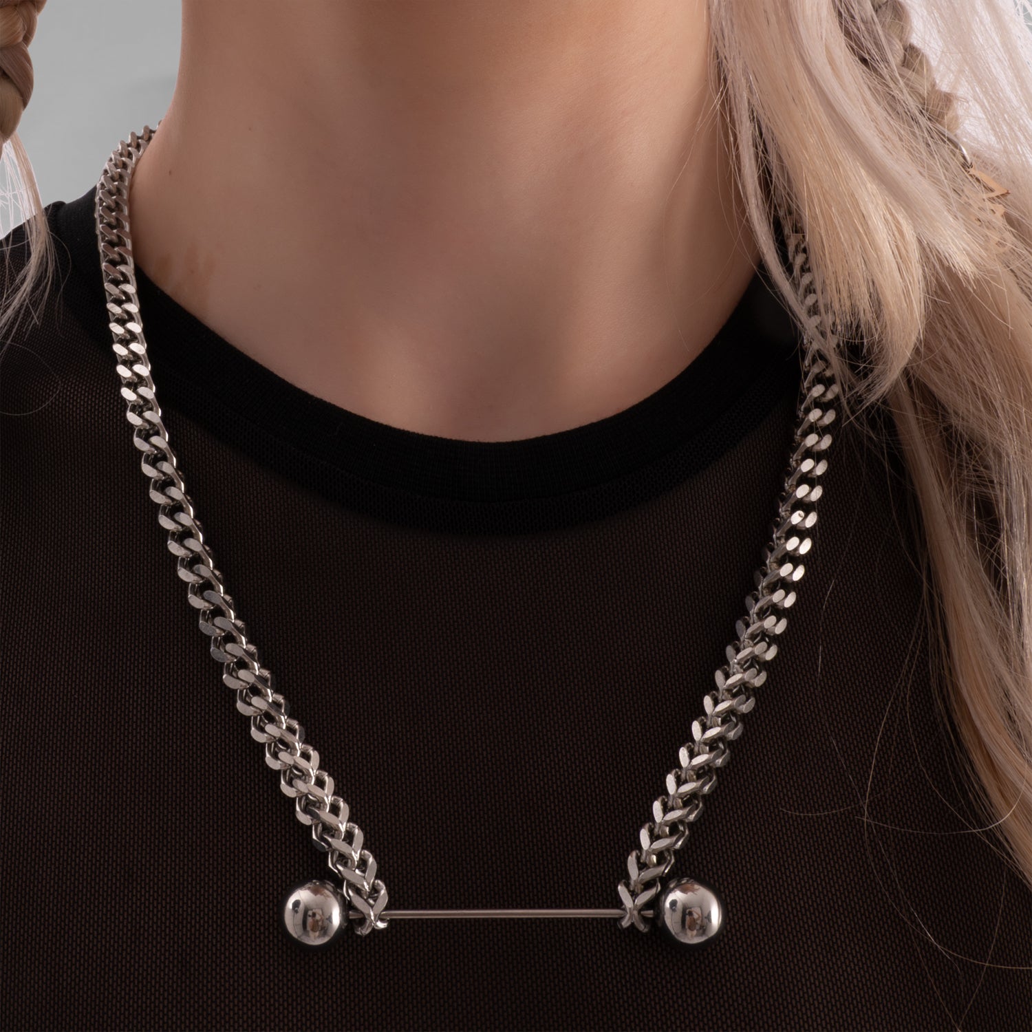 Close-up of a woman wearing a bold silver chain necklace with industrial barbell detail, styled with a black mesh top and platinum blonde braided hair, featuring facial piercings and red lipstick.