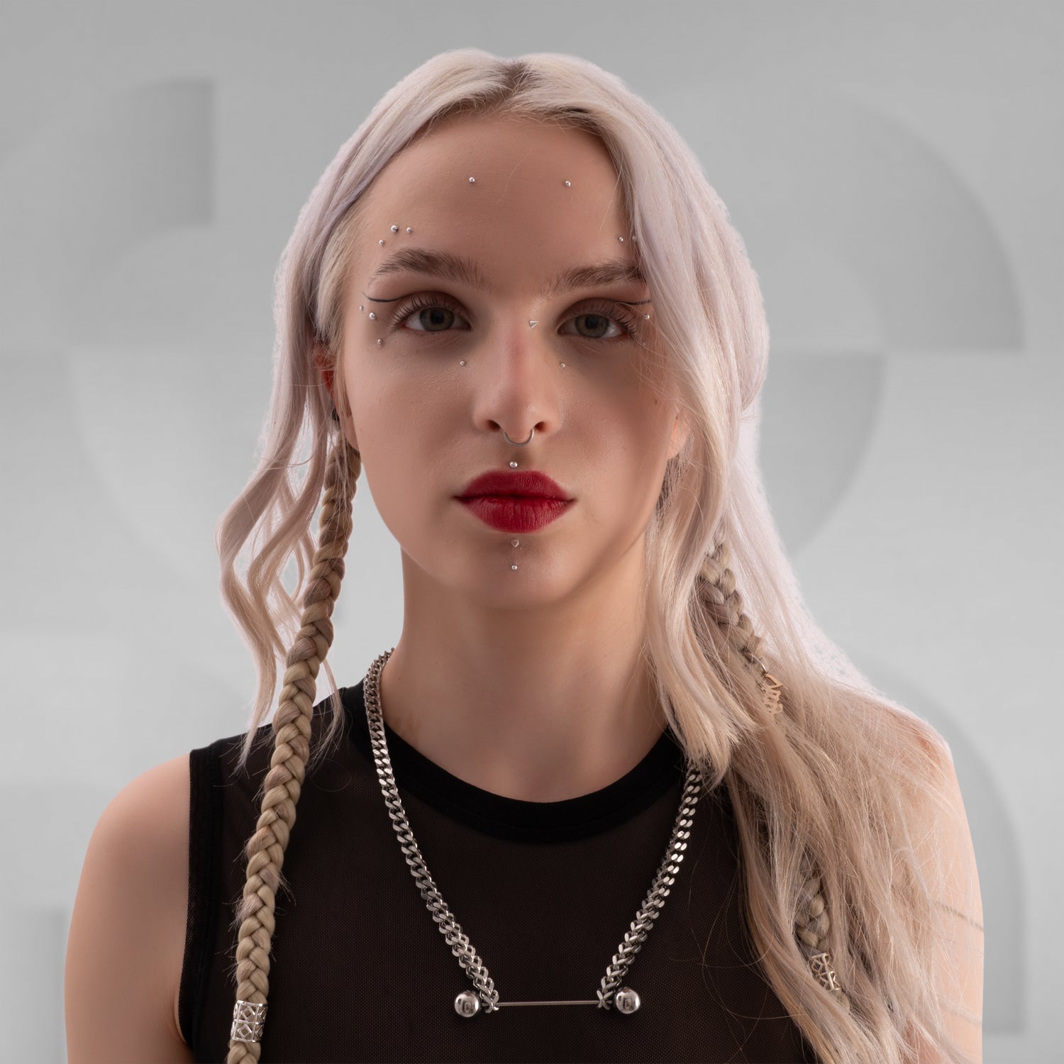 Close-up of a woman wearing a bold silver chain necklace with industrial barbell detail, styled with a black mesh top and platinum blonde braided hair, featuring facial piercings and red lipstick.