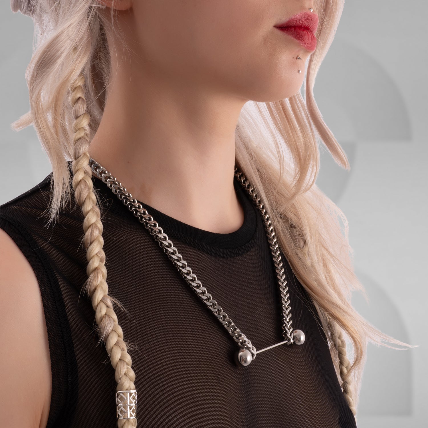 Close-up of a woman wearing a bold silver chain necklace with industrial barbell detail, styled with a black mesh top and platinum blonde braided hair, featuring facial piercings and red lipstick.