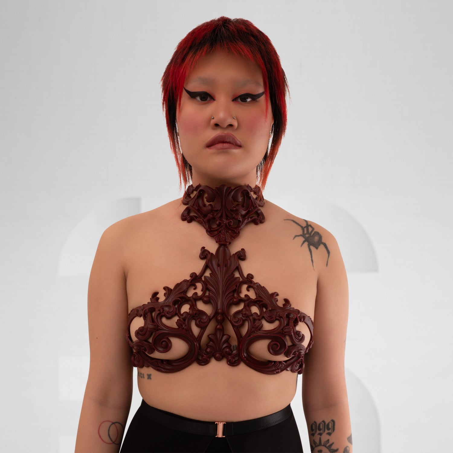 A trans model wearing a Baroque 3D Latex Bra in Wine, showcasing its elegant design. The bra features intricate detailing and complements the model's look against a light grey background, highlighting both style and individuality.