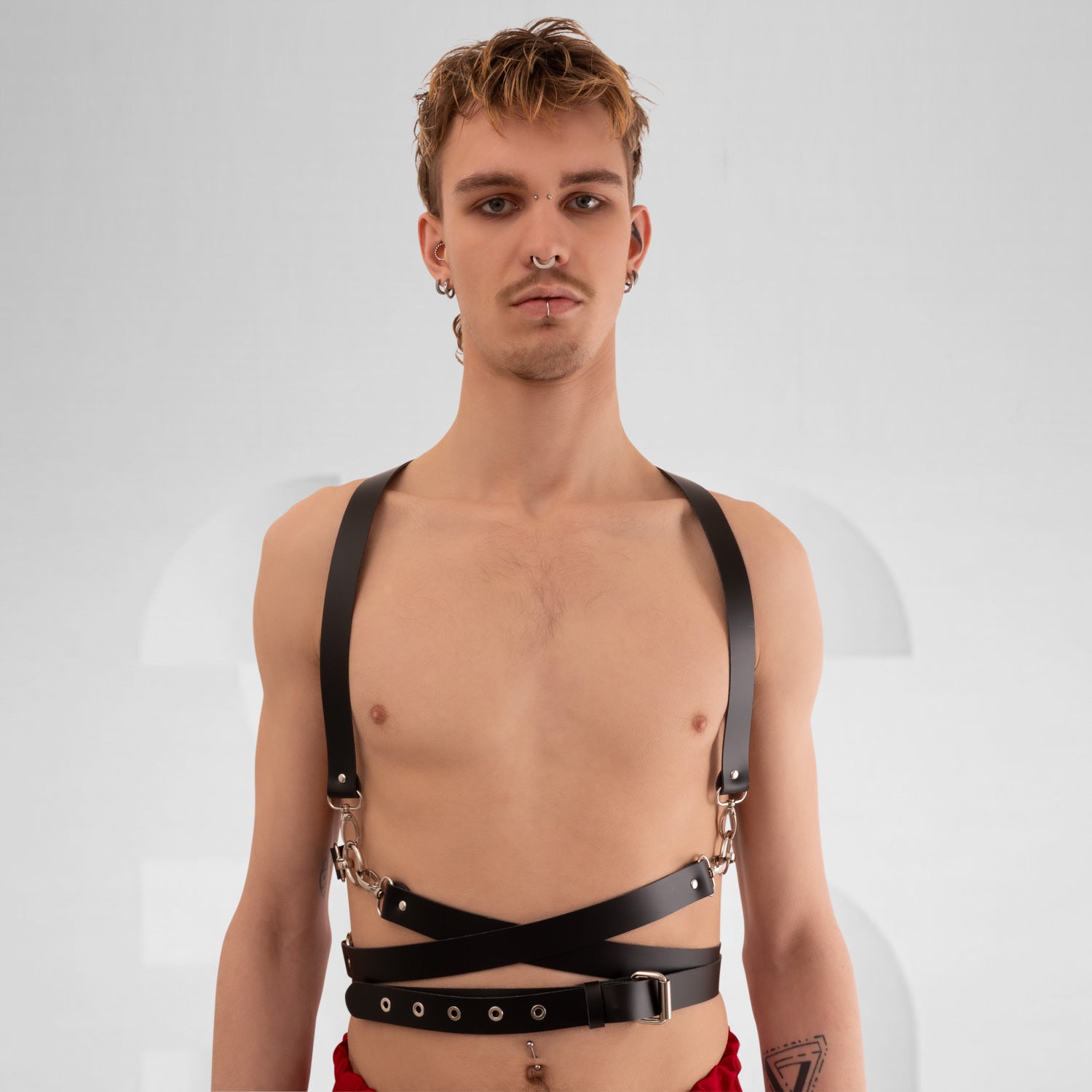  a stylish black leather harness with crisscross straps and metal hardware, creating a bold and modern look. The minimalist design is complemented by the neutral background, highlighting the harness's unique features and the model's confident stance.