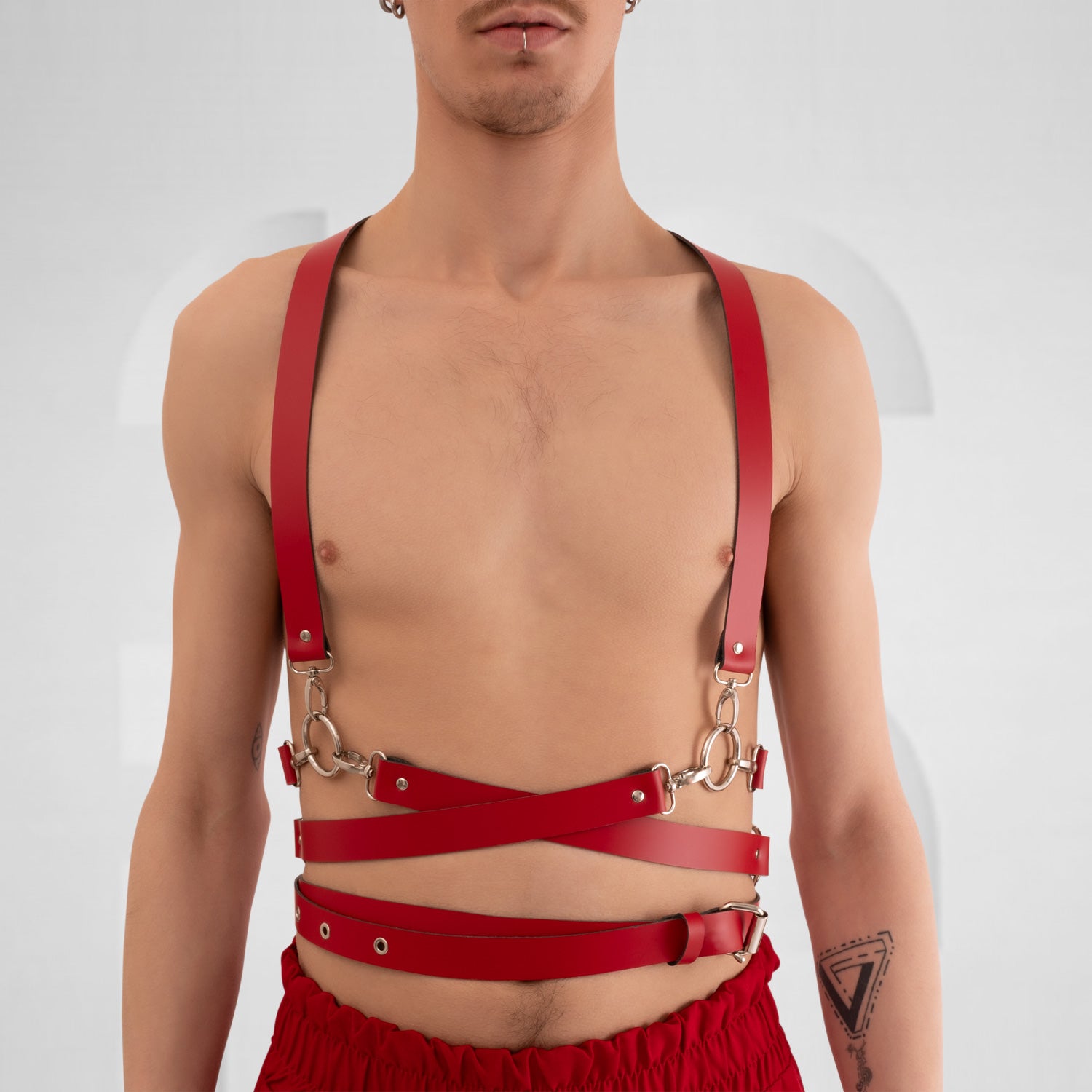  a stylish red leather harness with crisscross straps and metal hardware, creating a bold and modern look. The minimalist design is complemented by the neutral background, highlighting the harness's unique features and the model's confident stance.