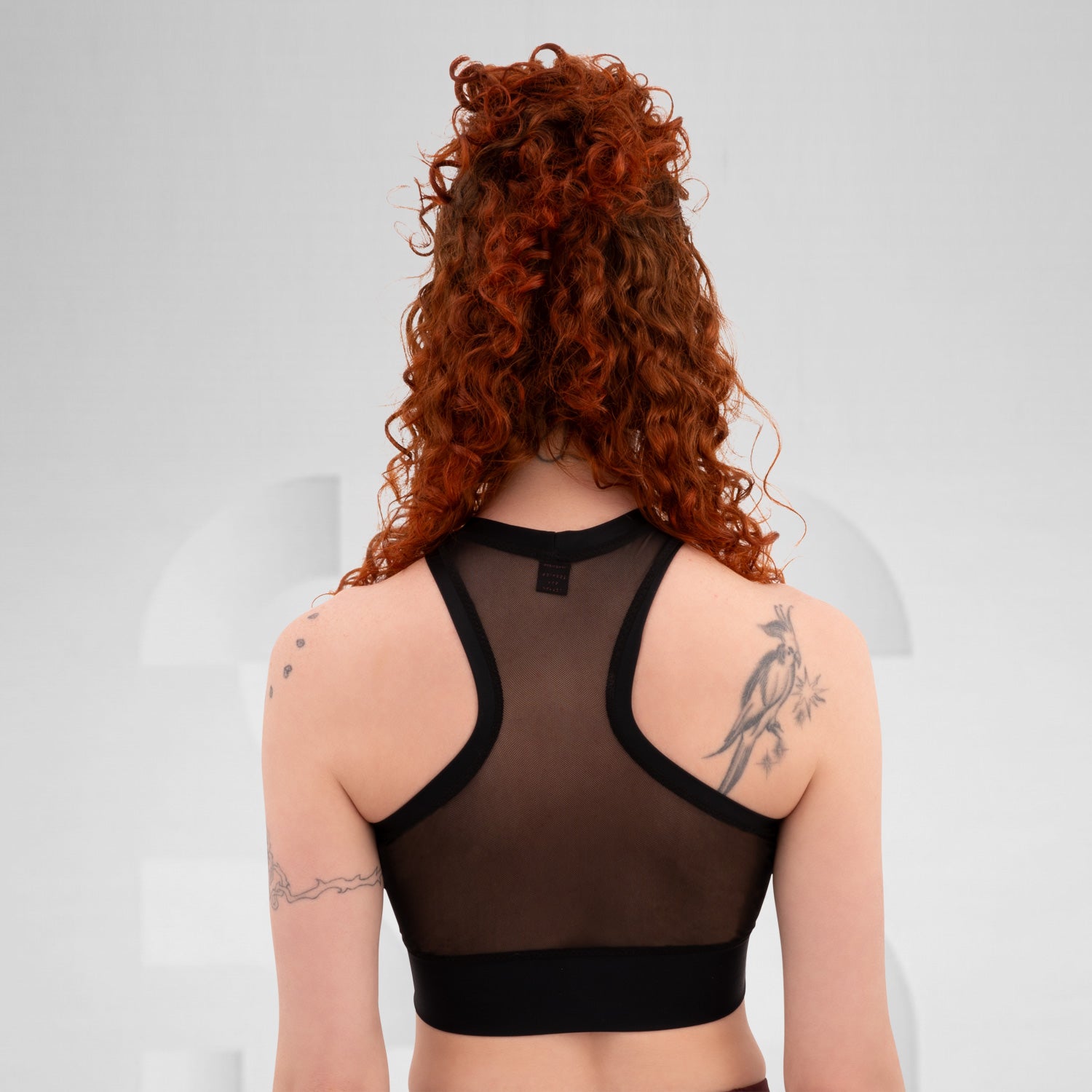 The a model wearing a black, sheer top with a sporty cut, featuring a layer of transparency. The outfit is accented by a chunky choker with a metallic detail. The model's arms are tattooed, showcasing various designs that add character to the look. The neutral background draws focus to the ensemble and the model's confident expression.