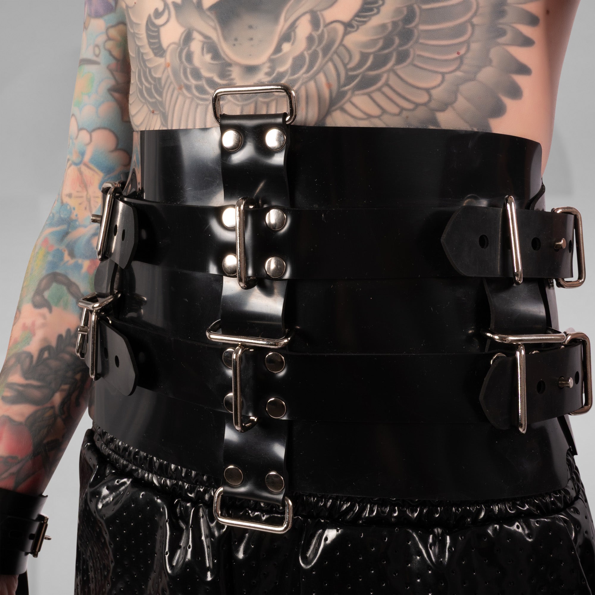 Black leather harness with metal buckles worn by a person with tattoos on a gray background