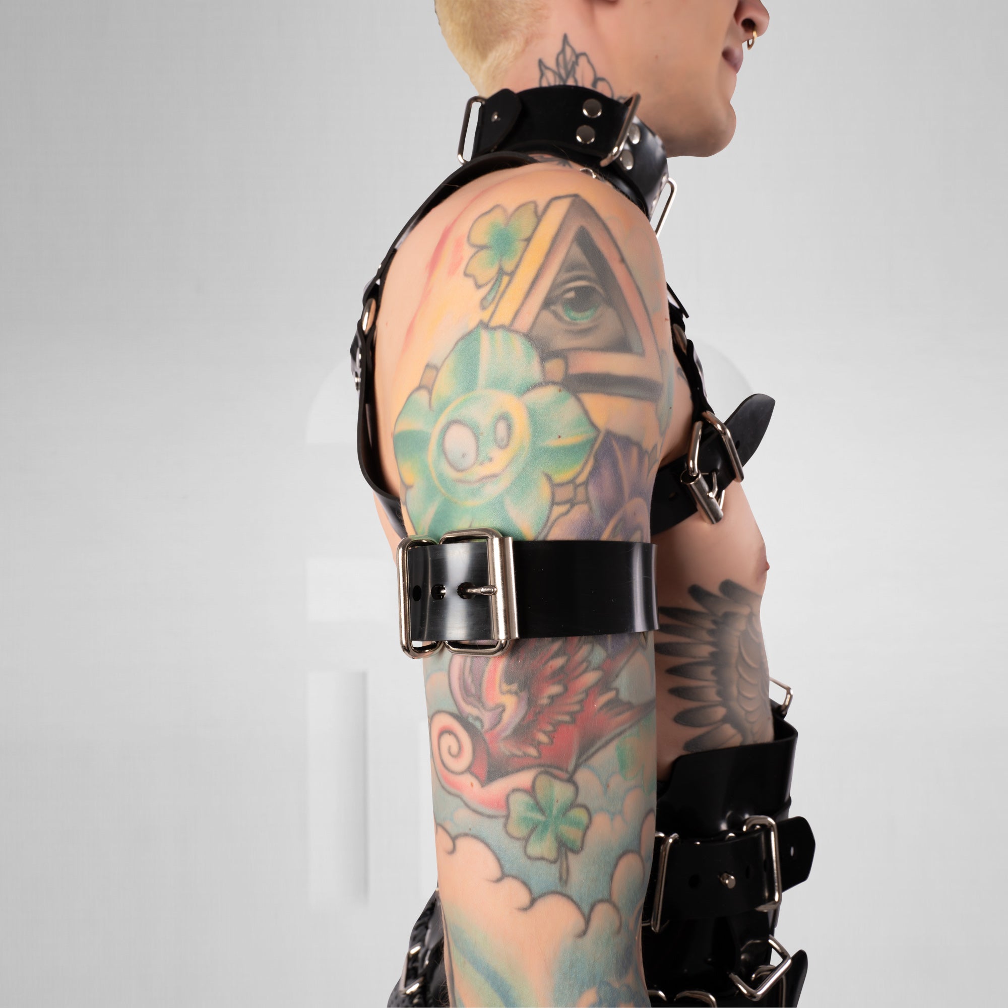 Person wearing a black harness with tattoos on arms and shoulders against a gray background