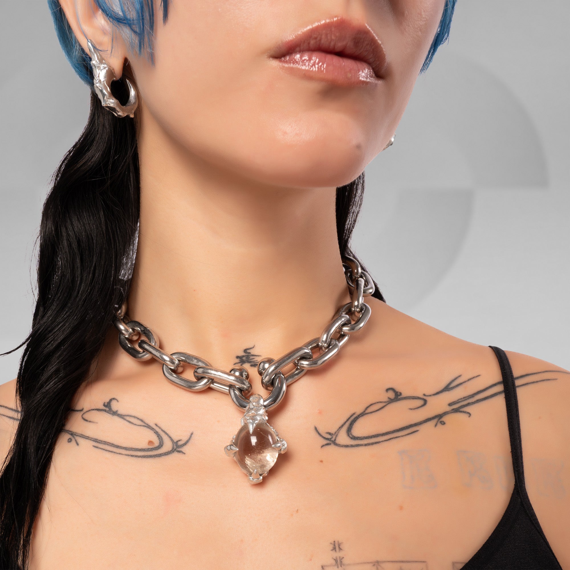 A close-up of a model wearing a chunky silver chain necklace with a clear faceted teardrop crystal pendant. The model has blue hair, black tattoos, and is wearing a black top against a neutral gray background.