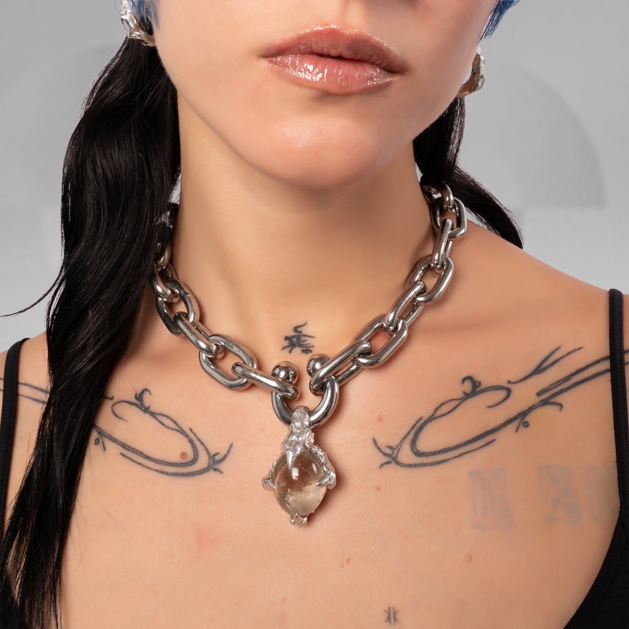 A close-up of a model wearing a chunky silver chain necklace with a clear teardrop-shaped crystal pendant, shown against her bare shoulders.