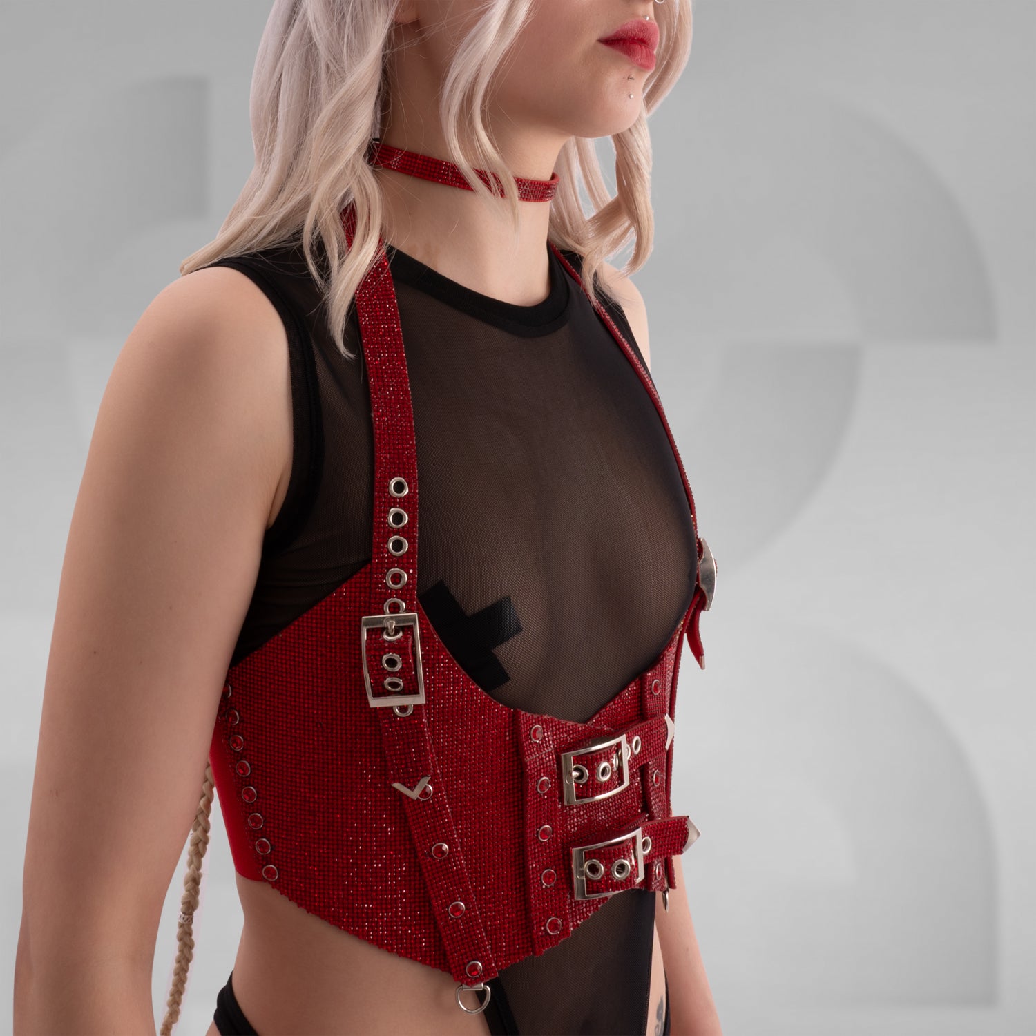 This image shows a person wearing a fashion outfit that includes a sheer black top and a red harness or corset-style accessory with buckle details. The accessory is made of a textured, glittery material. Additionally, the person is wearing a matching red choker. This style is edgy and modern, often seen in alternative fashion scenes.