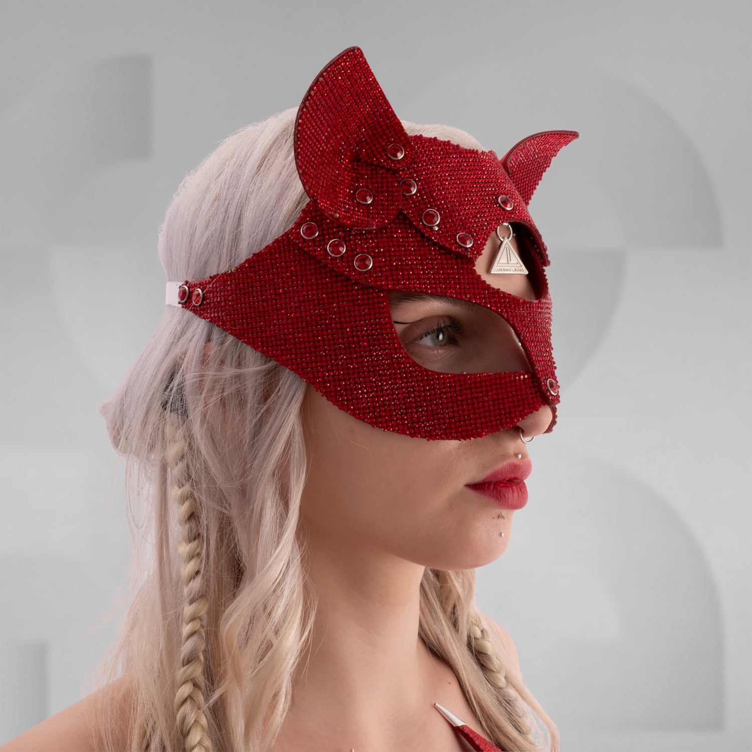 Woman wearing a sparkling rhinestone cat mask with pointed ears, paired with a matching crystal choker and shimmering outfit; styled with platinum blonde hair in braids and bold red lipstick, set against a minimalist grey background.