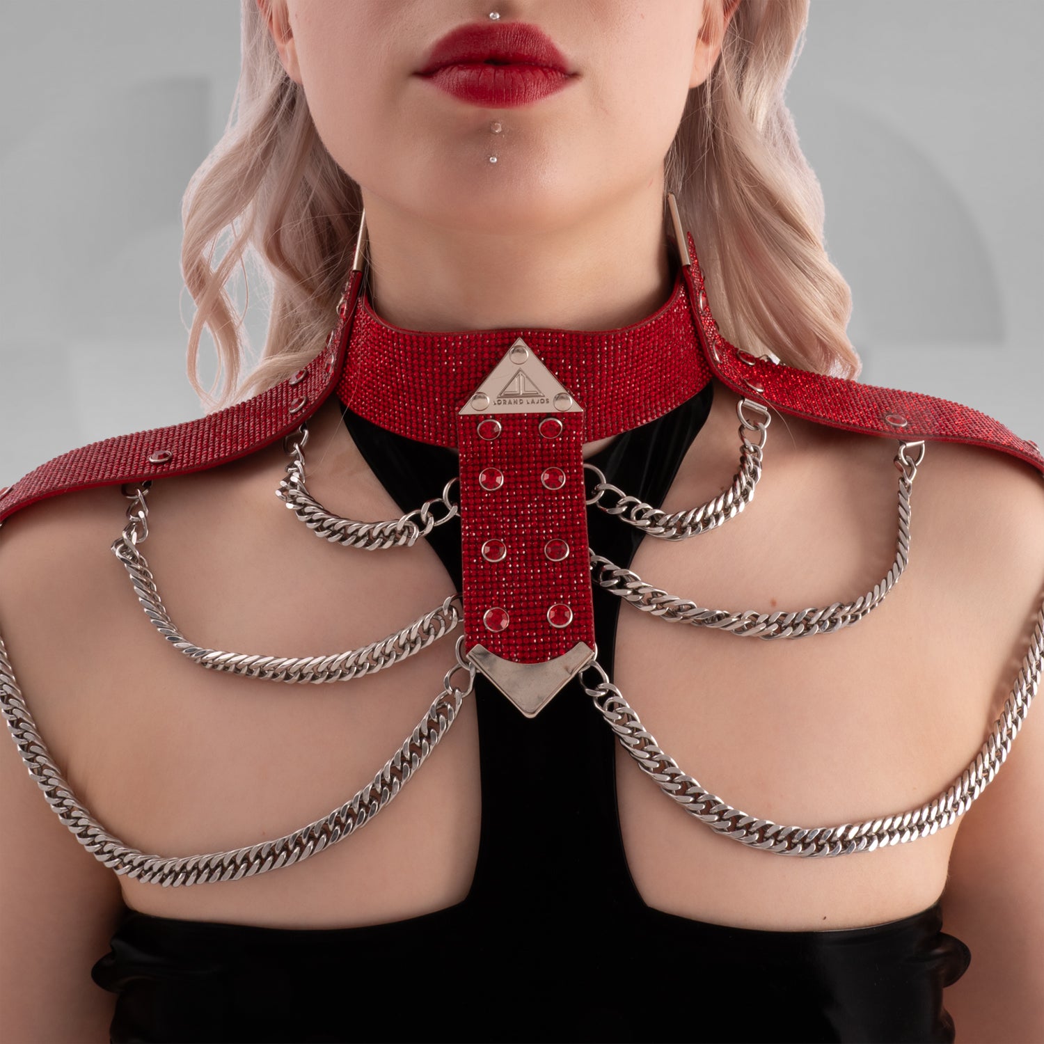 Close-up image of a person wearing a bold, red rhinestone-studded shoulder harness with silver chain details and a triangular metal centerpiece, styled over a black outfit—perfect for statement fashion or kinky-inspired looks.