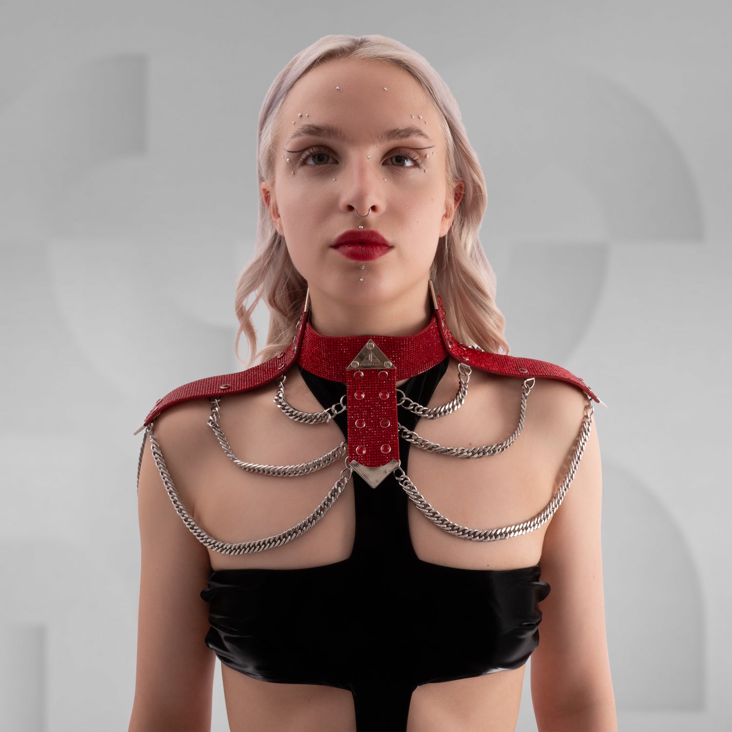 Close-up image of a person wearing a bold, red rhinestone-studded shoulder harness with silver chain details and a triangular metal centerpiece, styled over a black outfit—perfect for statement fashion or kinky-inspired looks.