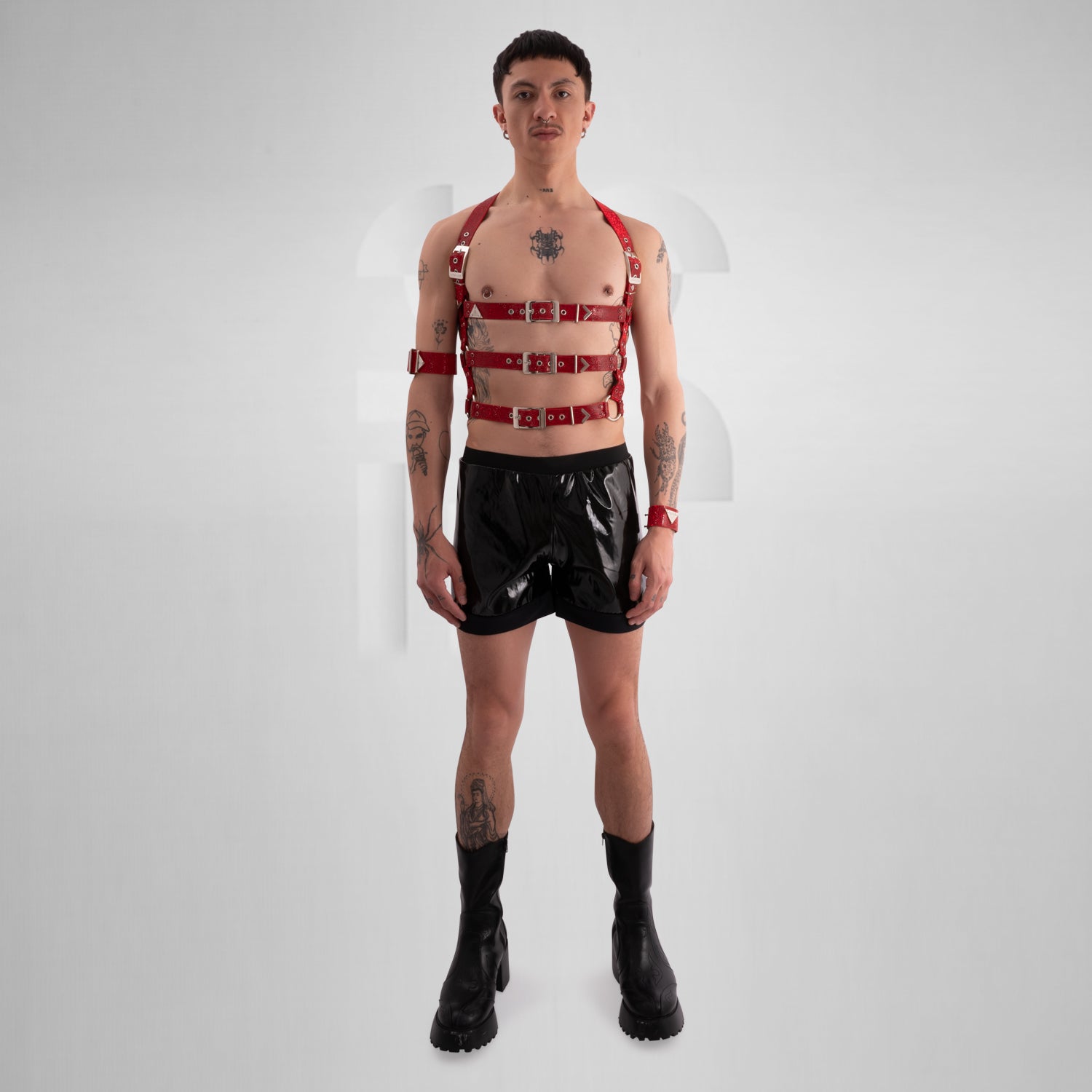 This image showcases a person with a confidently rebellious style, featuring multiple tattoos on their chest, arms, and neck. They have short, dark hair and a septum piercing. The individual is wearing a bold, black harness made of sturdy, glittering material with multiple adjustable straps and silver hardware, which encases the chest and shoulders. Their makeup is minimal, emphasizing their natural features. 